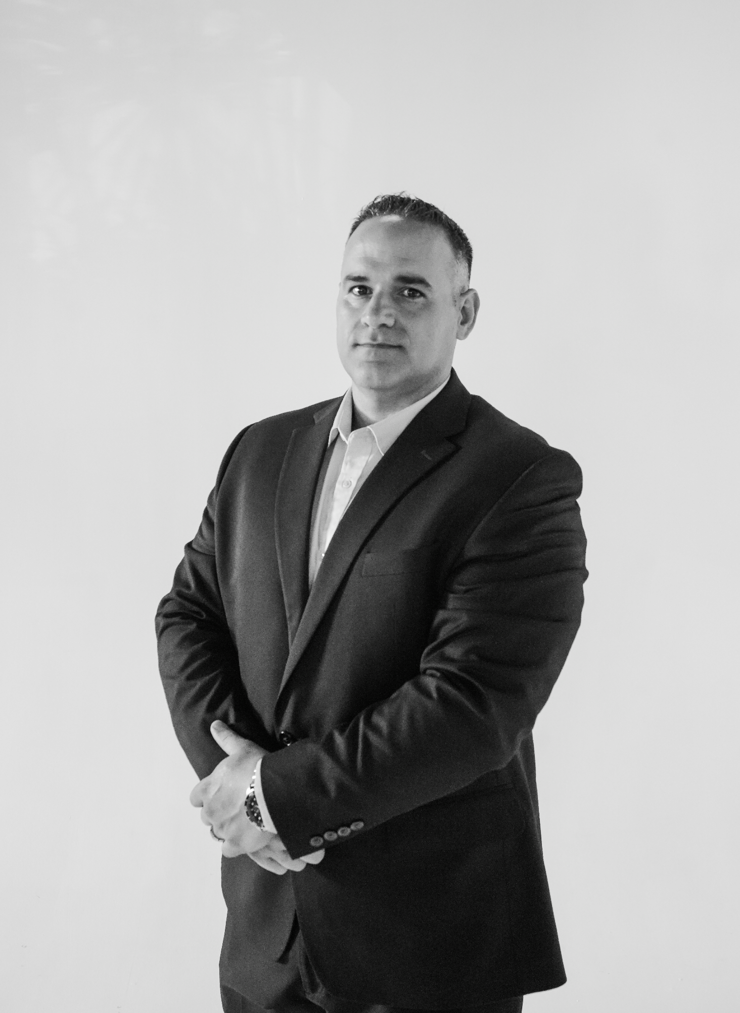 A man in formal attire standing against a white background, looking at the camera with arms crossed.
