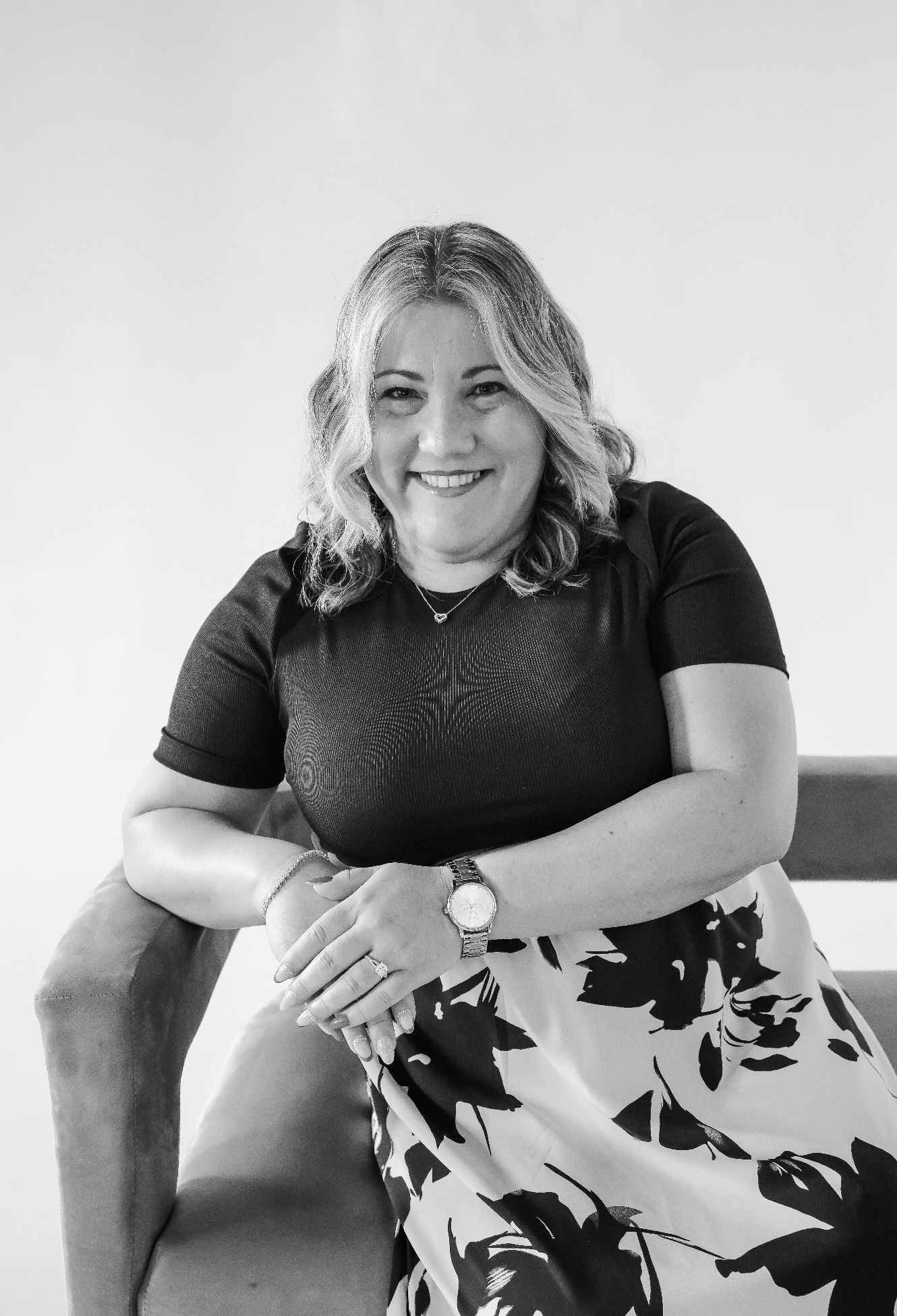 A woman with shoulder-length wavy hair, smiling, wearing a black top, floral skirt, and watch, sitting on a chair with her arms resting on the armrest, in black and white.