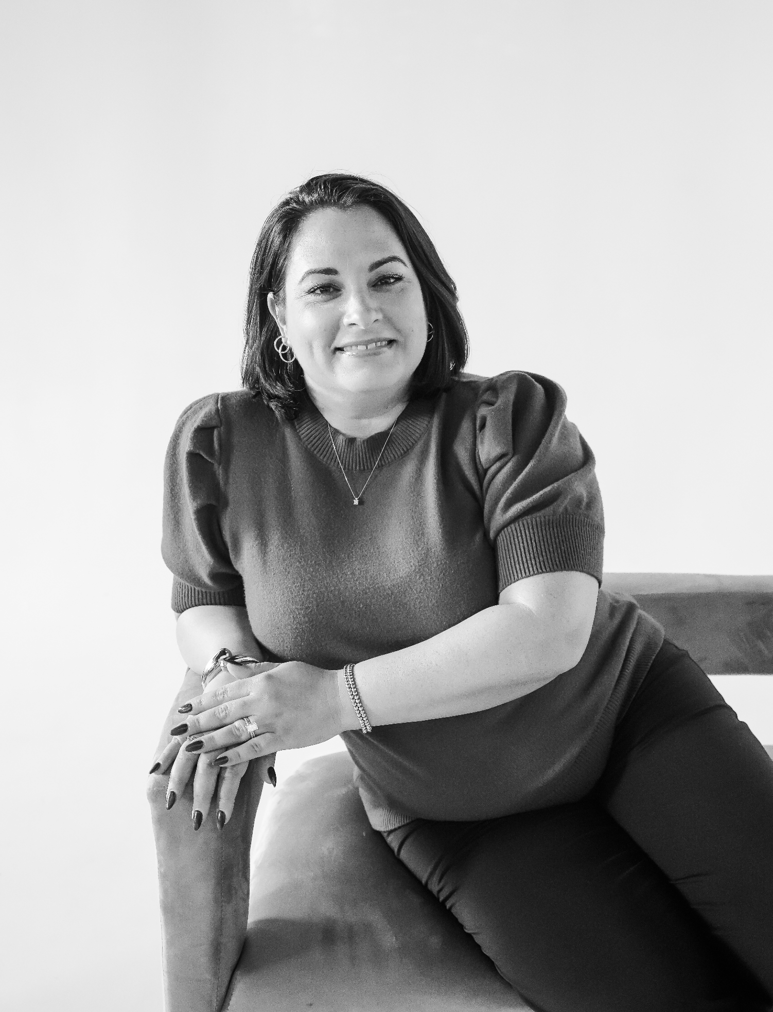 Black and white photo of a woman with shoulder-length dark hair, sitting on a chair, wearing a short-sleeved sweater and dark pants, smiling and looking at the camera.