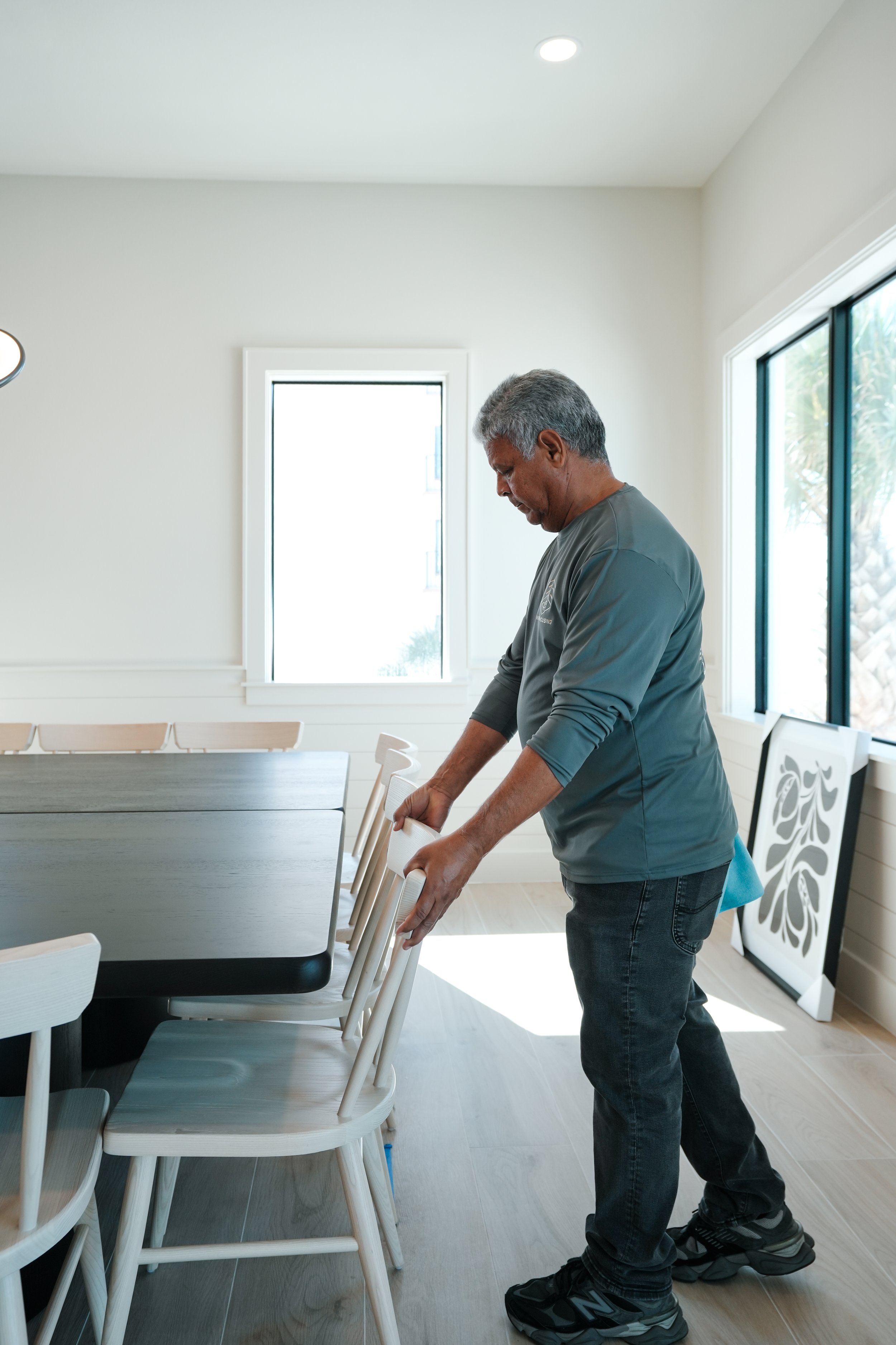 An older man in a gray long-sleeved shirt and black jeans is placing a white chair at a dining table in a bright room with light-colored walls and a wooden floor.