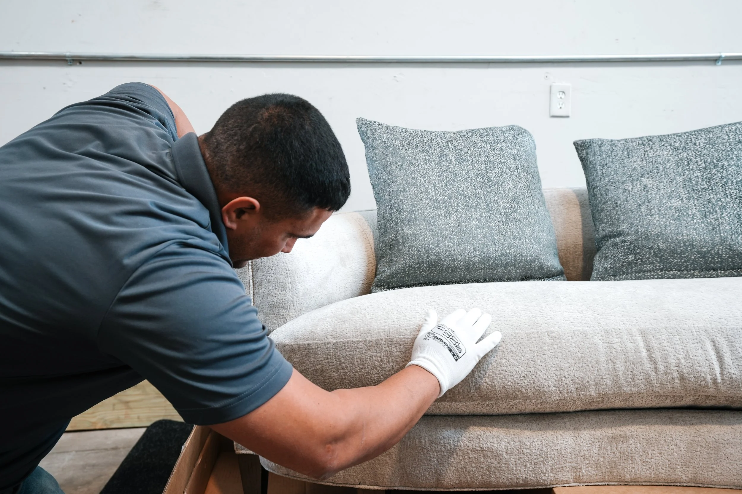 A man inspecting or repairing a beige fabric sofa with two gray patterned pillows, wearing a gray shirt and a white glove, inside a room with unfinished wooden floor and plain white walls.