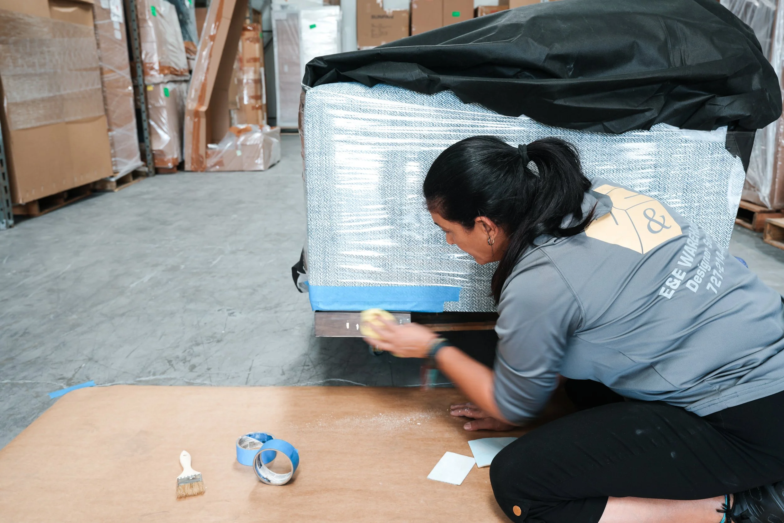 A woman working on wrapping or taping a large item covered in plastic and fabric in a warehouse with shelves filled with boxes in the background.