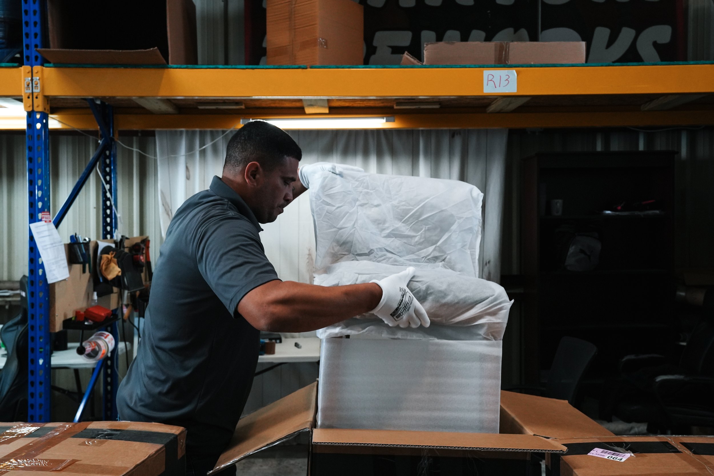 A man wearing a gray shirt and white gloves is handling a book inside a warehouse. He is standing at a table, opening a cardboard box with a large wrapped item inside. The background shows shelves with boxes and various tools or supplies.