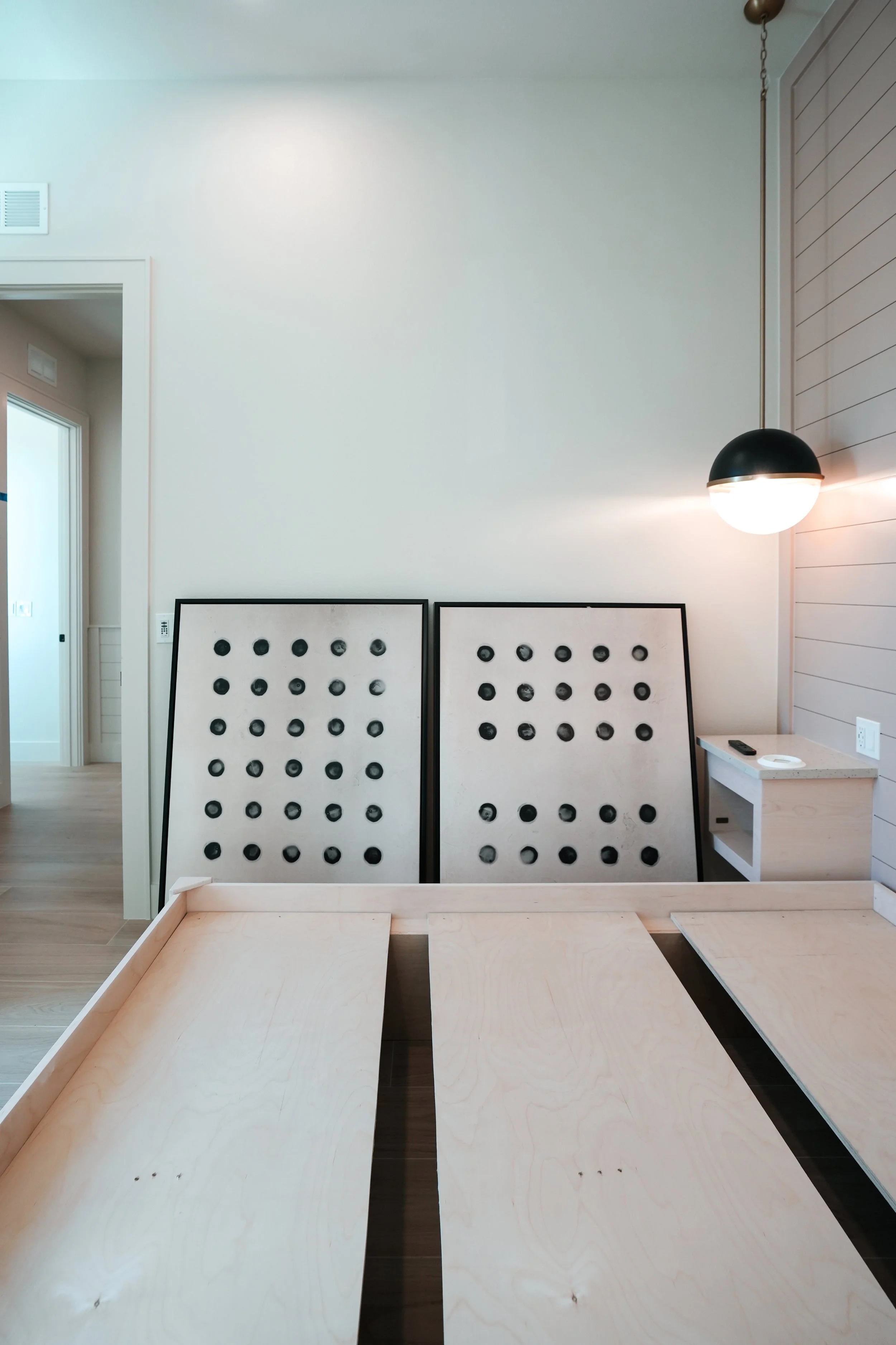 Interior of a modern bedroom showing two bed frames leaning against a white wall with black circular headboard accents, a small white side table, and a pendant light on the right side.