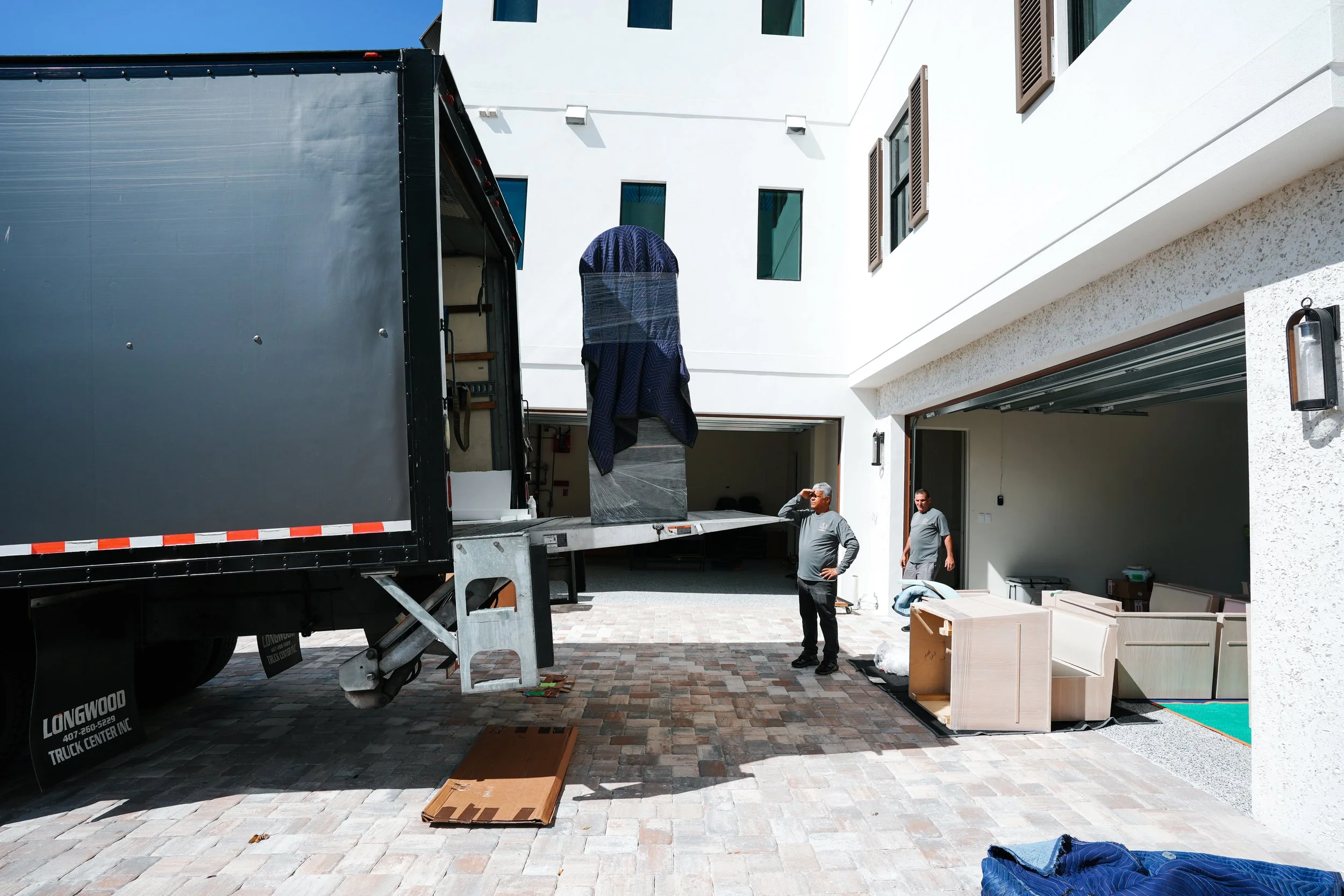 People unload furniture from a moving truck outside a modern white house with open garage doors.