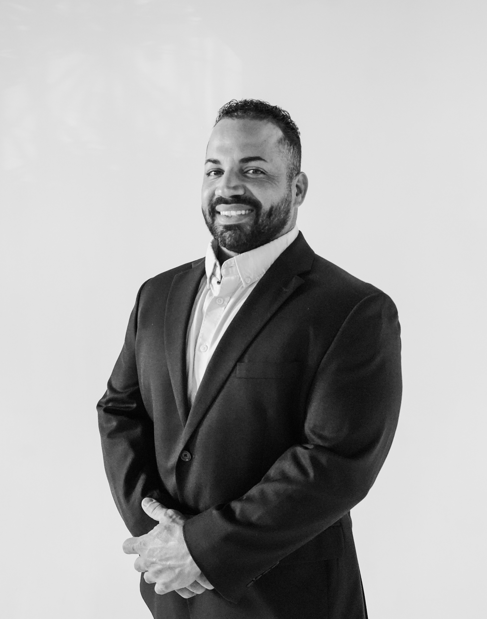 A man with a beard wearing a suit and white shirt, standing against a plain light-colored background, smiling and looking at the camera.