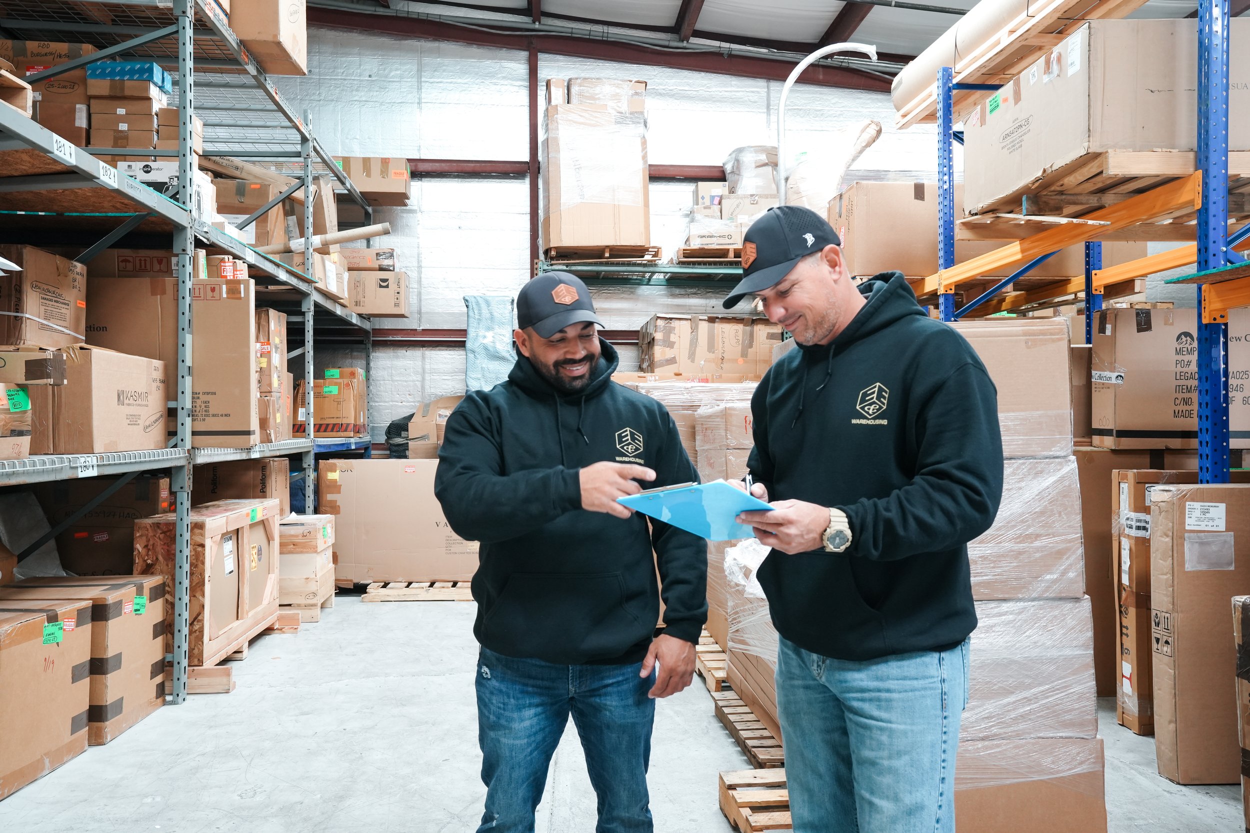 Two warehouse workers in black hoodies and caps looking at a clipboard and smiling inside a storage warehouse with shelves of boxes and pallets.