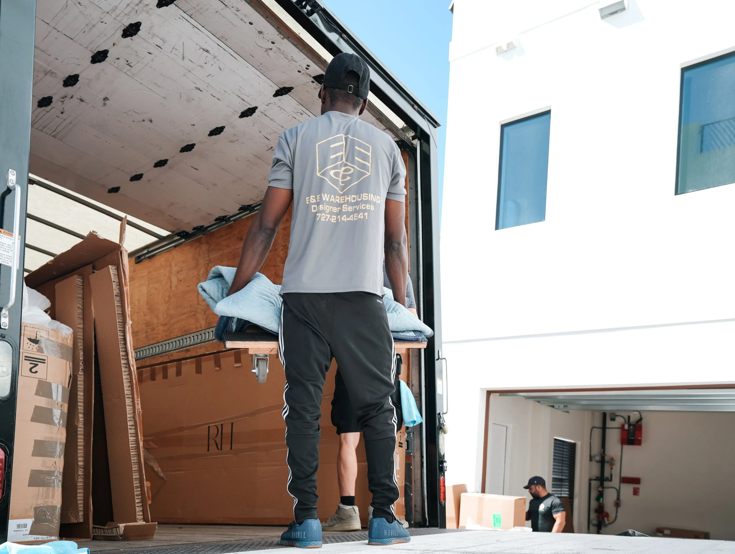 A man wearing a gray shirt and black pants is standing in a truck, carrying a blanket. The shirt has a logo and text that reads 'E&E Warehousing D Signer Services' with a phone number. There are cardboard boxes inside the truck. Another man is in the background near the garage, wearing a black shirt and cap, handling boxes.
