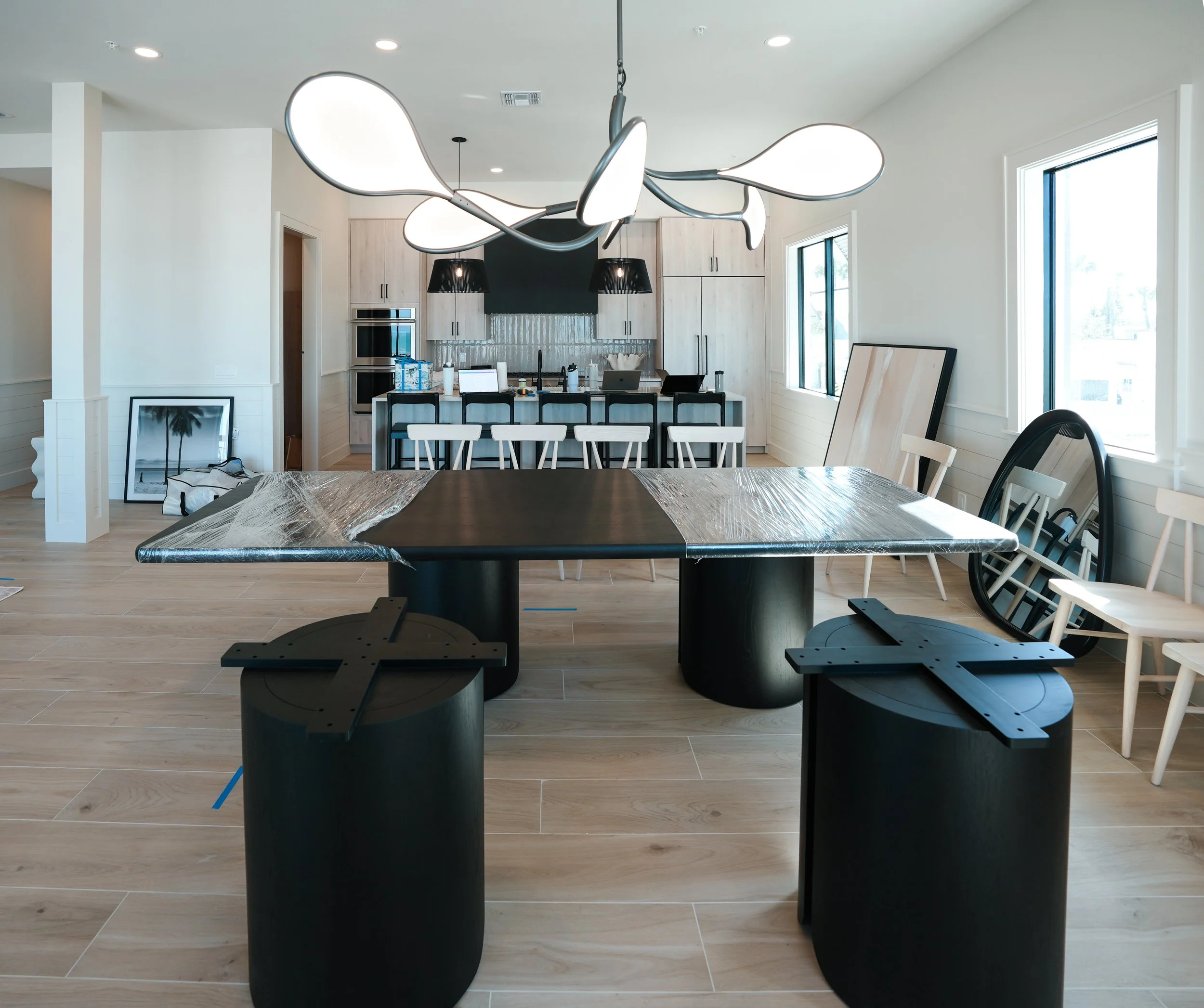 Modern kitchen and dining area with a table covered in plastic wrap, surrounded by black stools and white chairs, featuring large windows and contemporary lighting. In a new, custom build home that is in the greater Tampa Bay area.