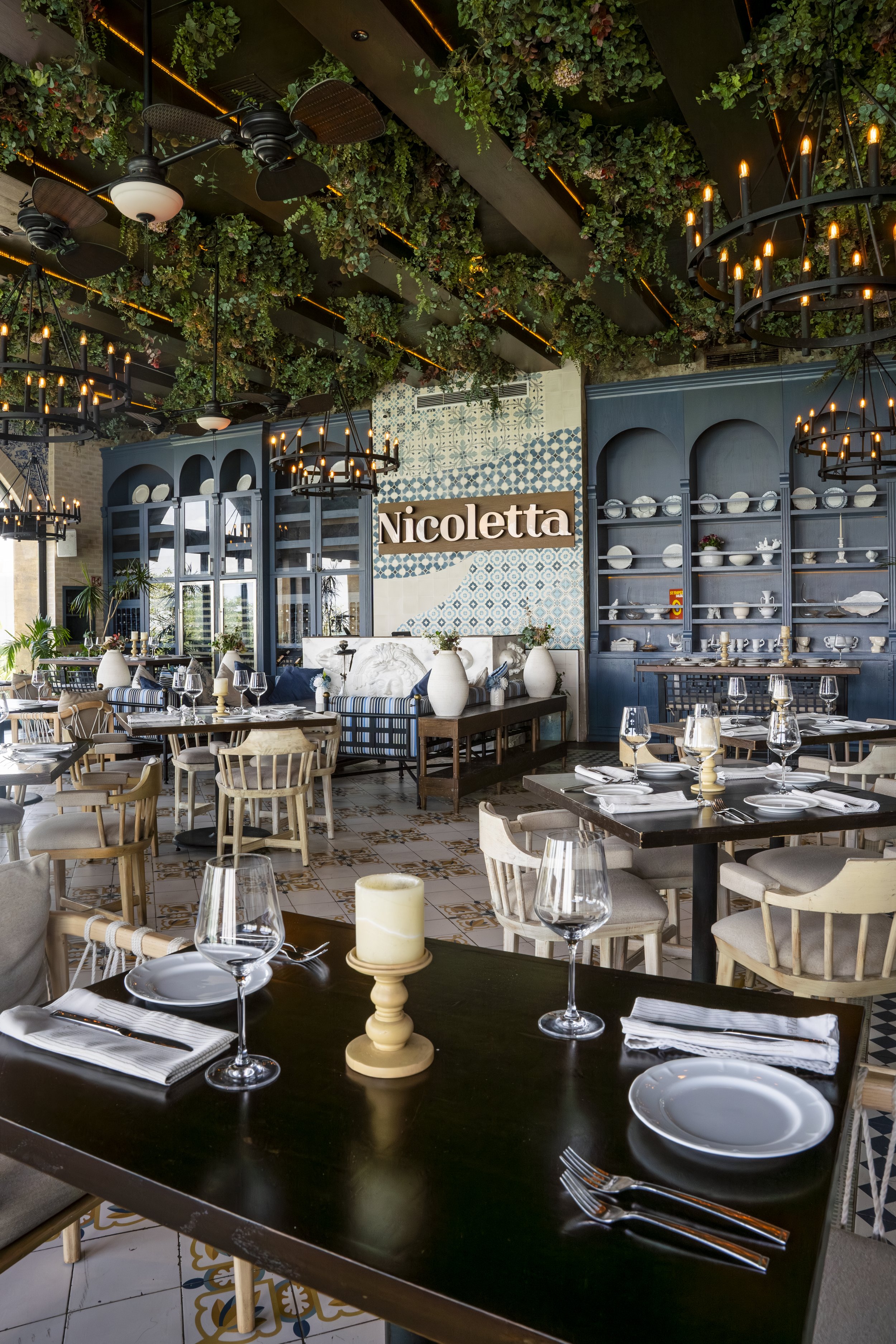 Inside a restaurant with a ceiling decorated with greenery, large black chandeliers, and ceiling fans. There are many tables set with wine glasses, plates, and cutlery. A sign reads 'Nicoletta' on a tiled wall with blue cabinets displaying dishes and decor.