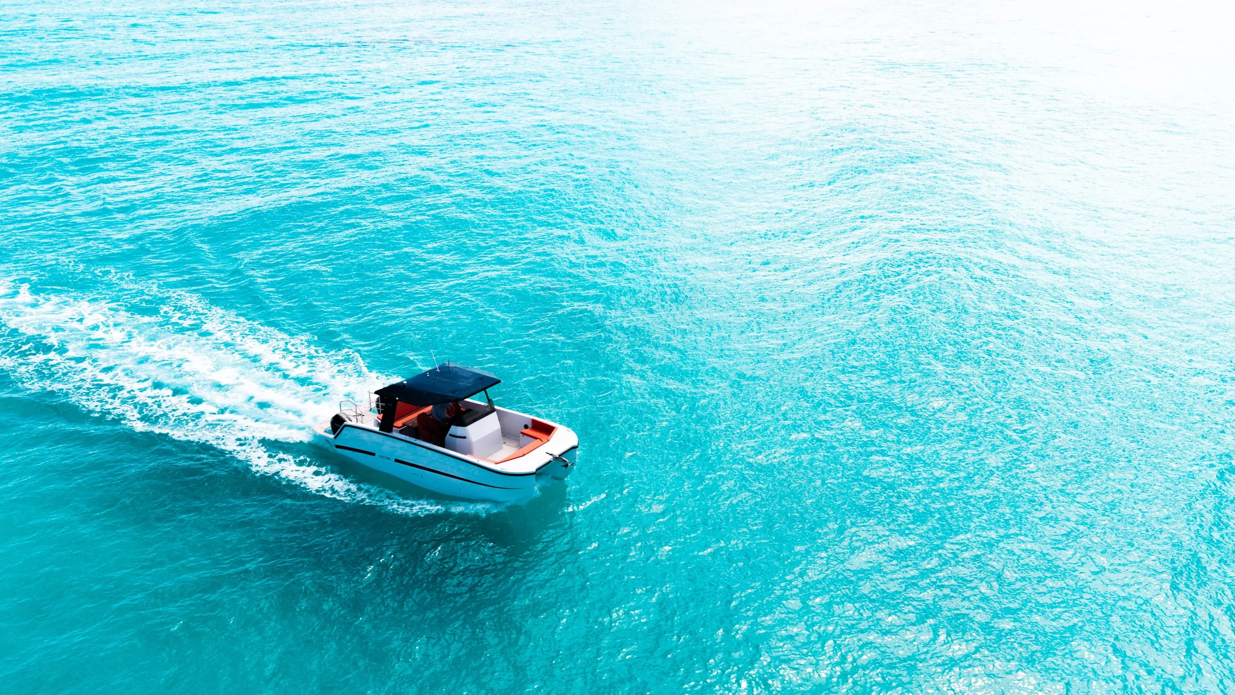 A small white boat with a black roof seen on the water, creating a wake as it moves through the calm, turquoise ocean.