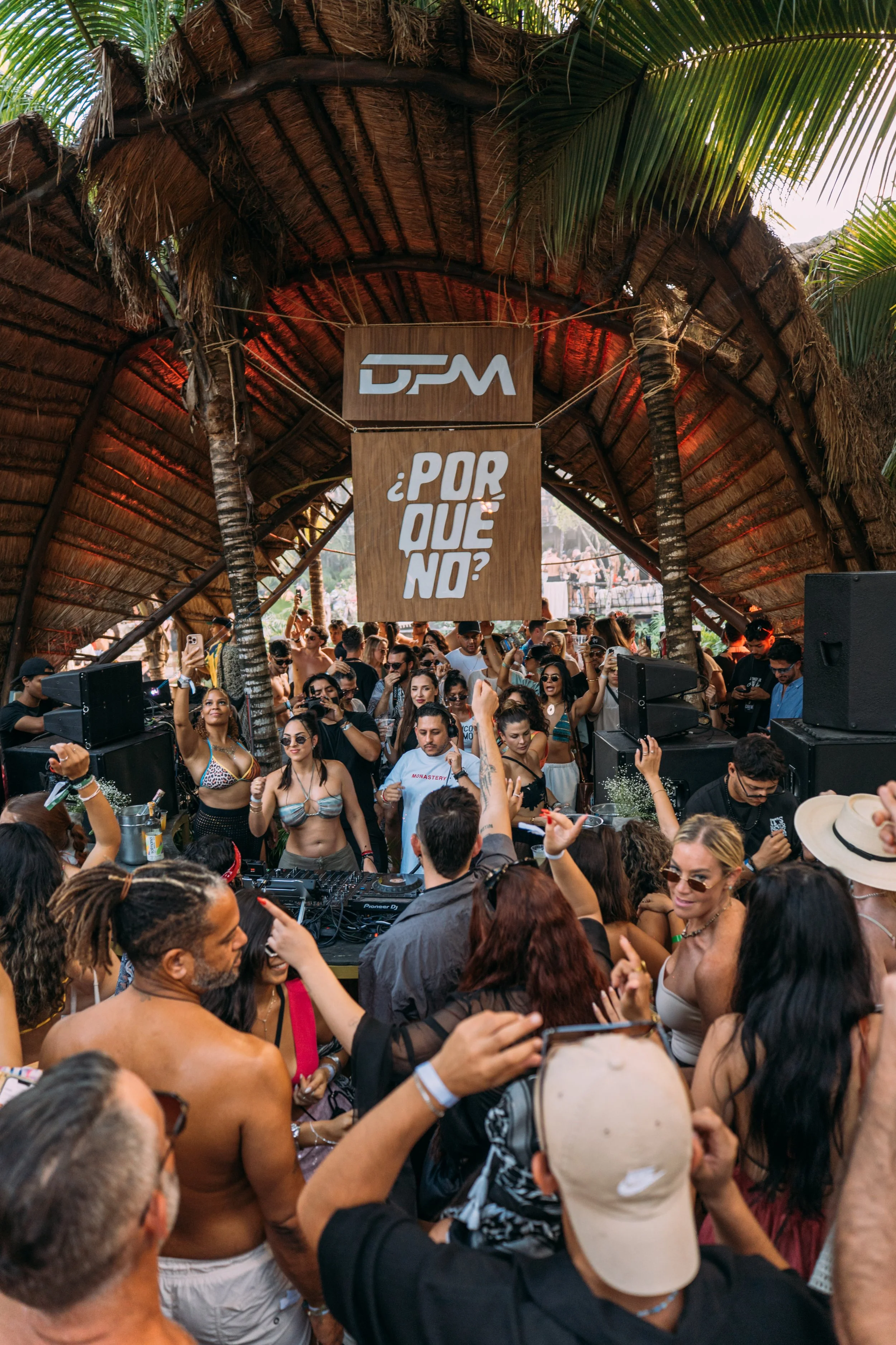 A lively outdoor DJ event held under a thatched roof with palm trees. The crowd is dancing and enjoying music, with some individuals raising their hands. There are large speakers and a DJ booth in the center.