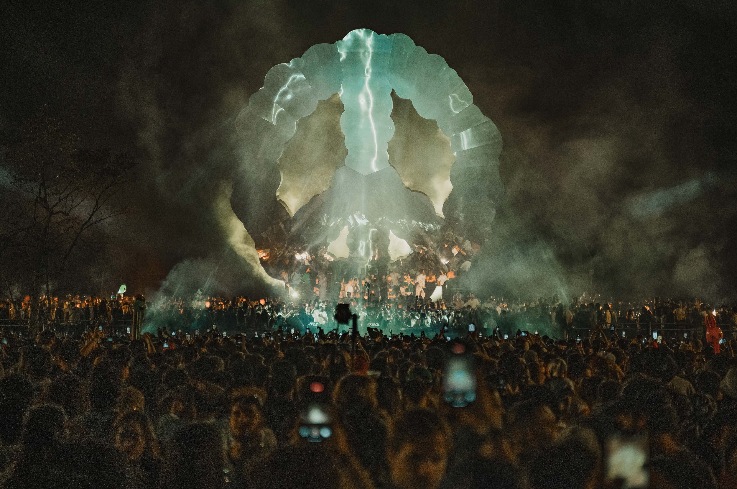 A large crowd attending a nighttime event with a bright, illuminated arch-shaped structure resembling a peace symbol and smoke in the background.