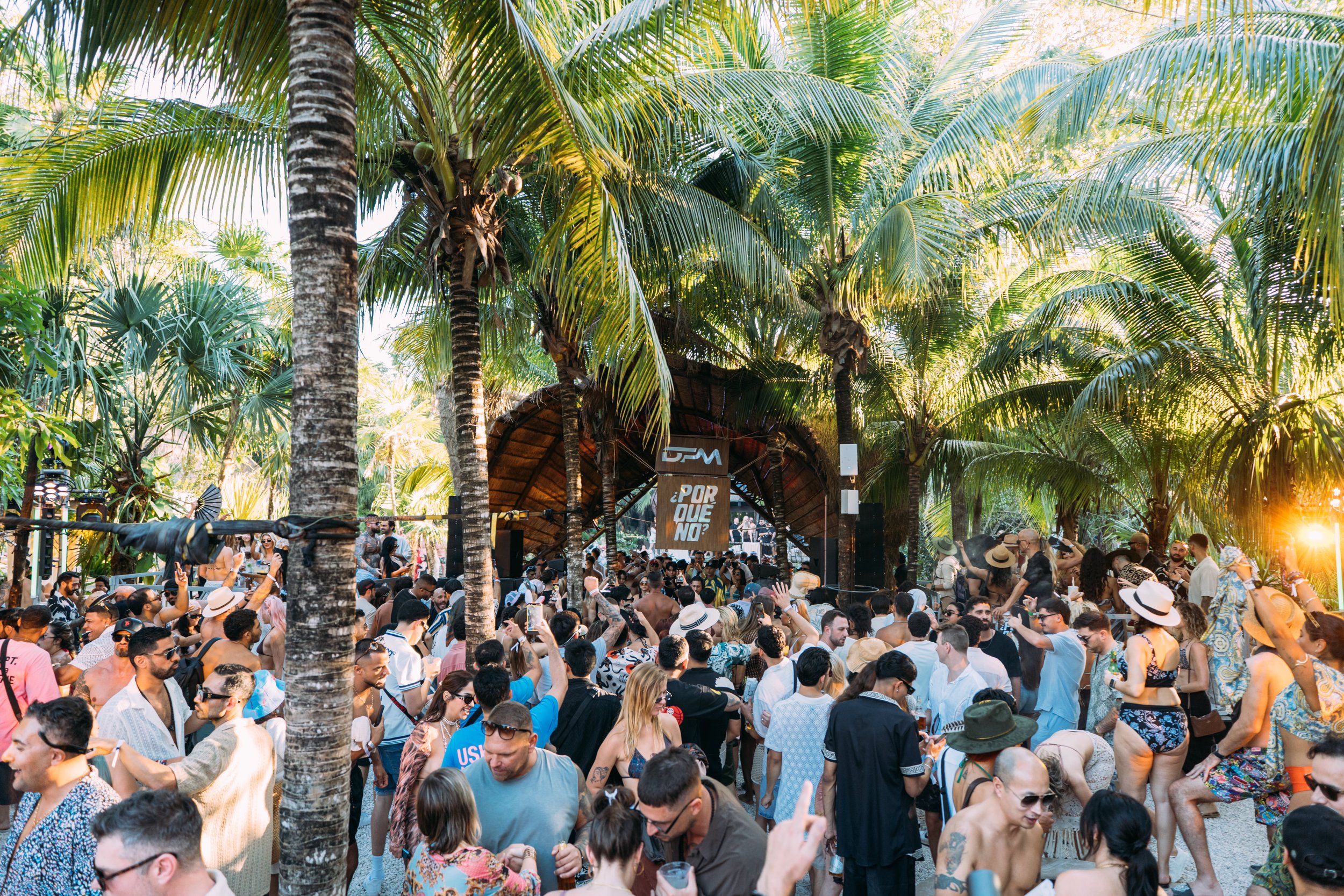 Crowd of people at an outdoor tropical music festival with palm trees, sun setting, and stage in the background.