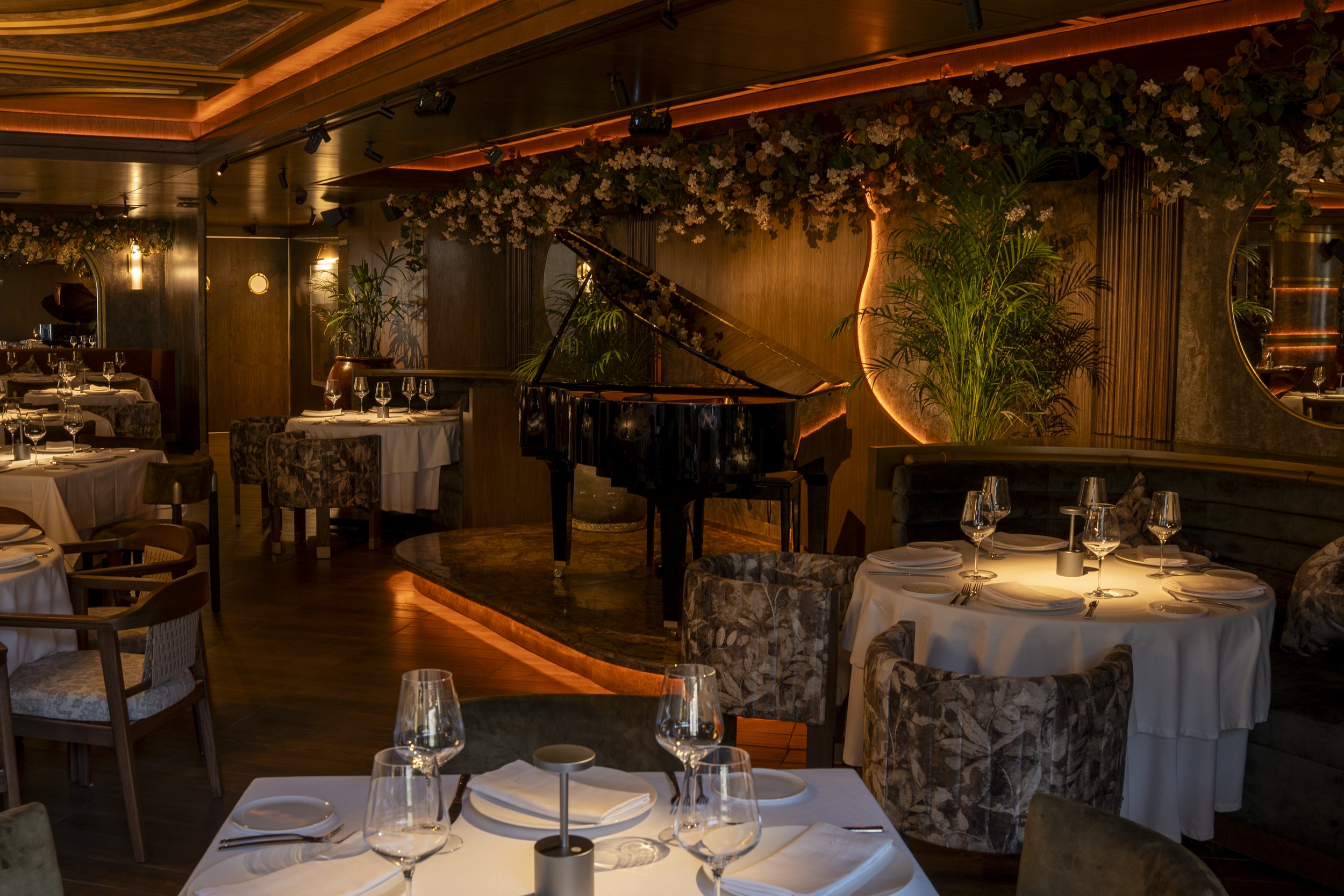 Elegant restaurant interior with a grand piano on a small stage, surrounded by potted plants and decorated with floral arrangements, dim lighting, and set tables with white linens and glassware.