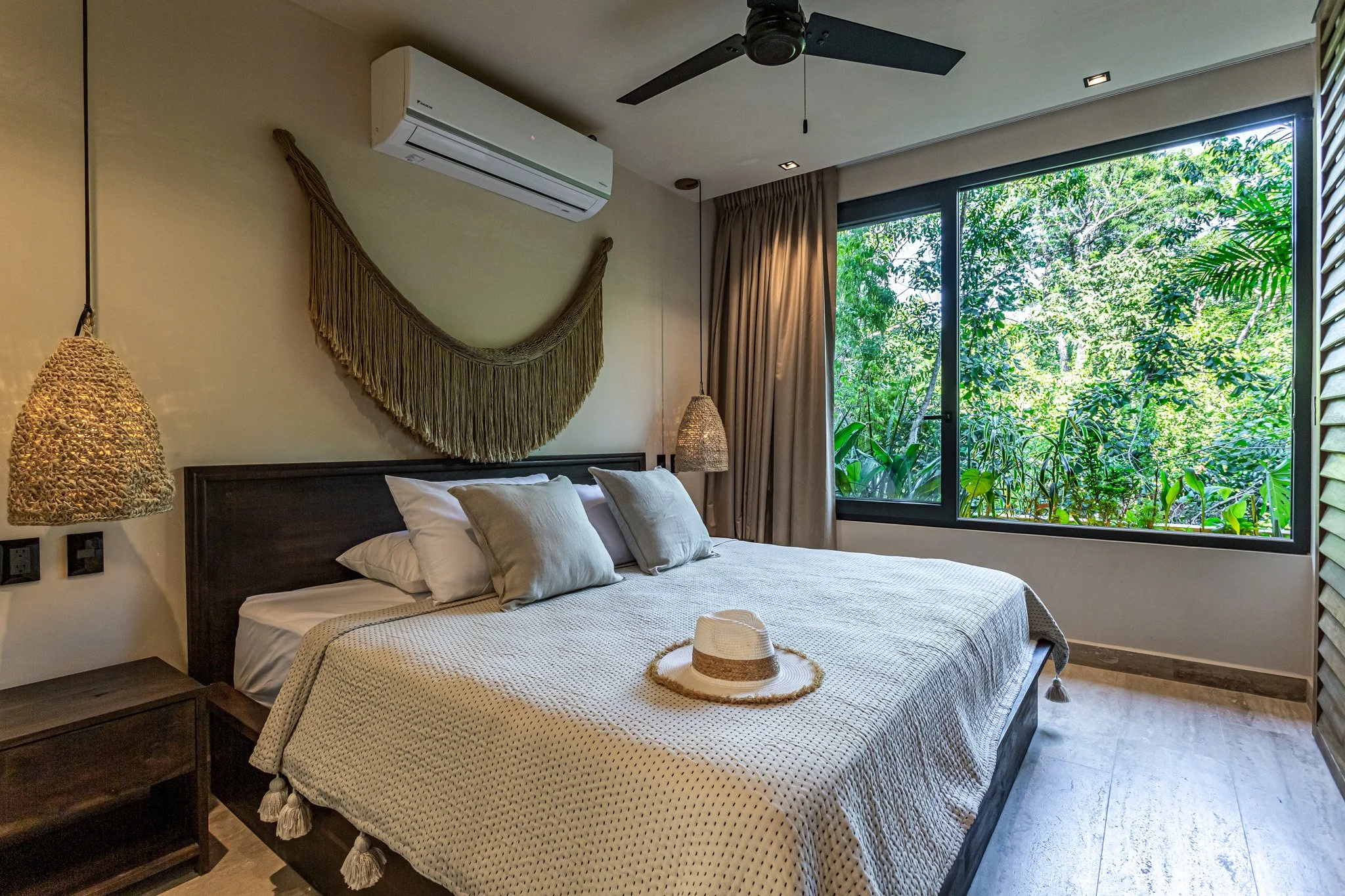 Bedroom with a large window showing lush green trees, a bed with white and beige pillows, beige textured blanket, a straw hat on the bed, woven hanging lamps, a wall-mounted air conditioner, and beige curtains.