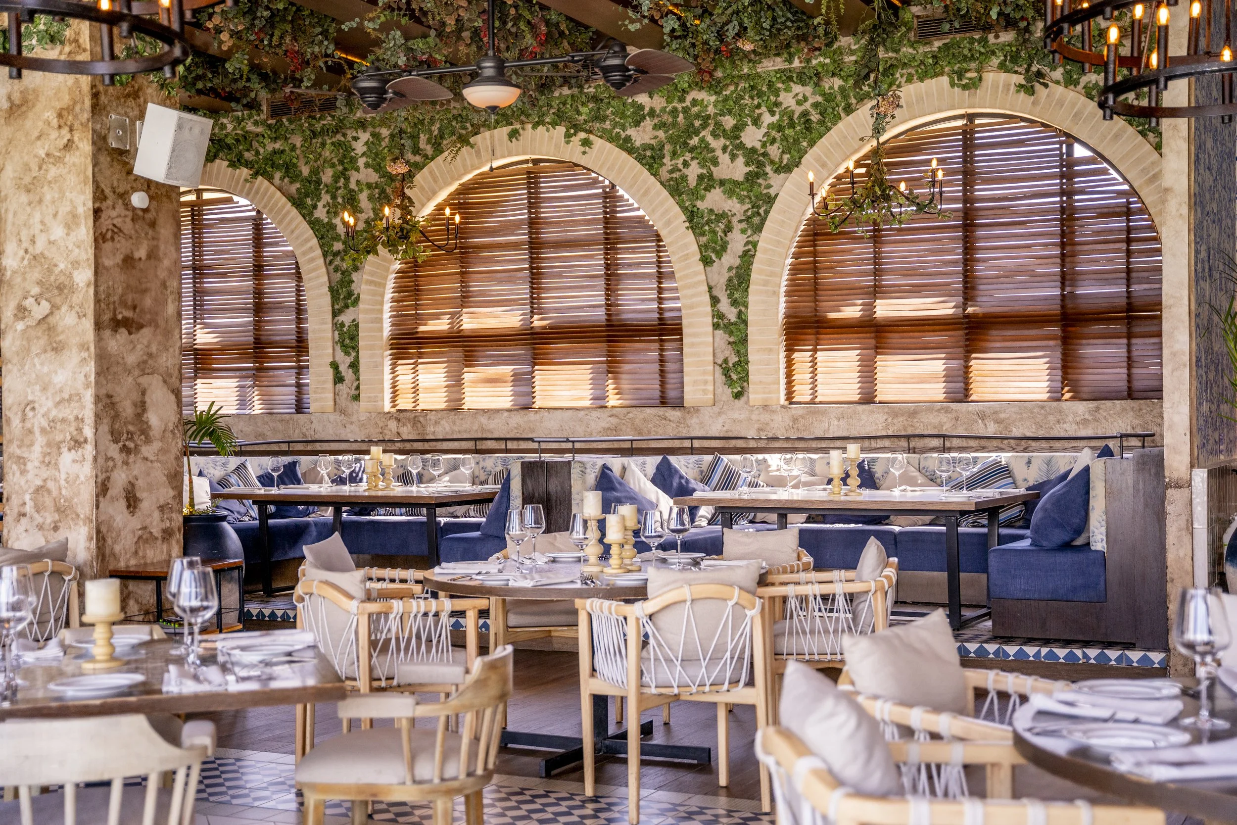 Interior of a restaurant with wooden furniture, blue and beige cushions, candles, and windows with wooden blinds, decorated with hanging plants and chandeliers.