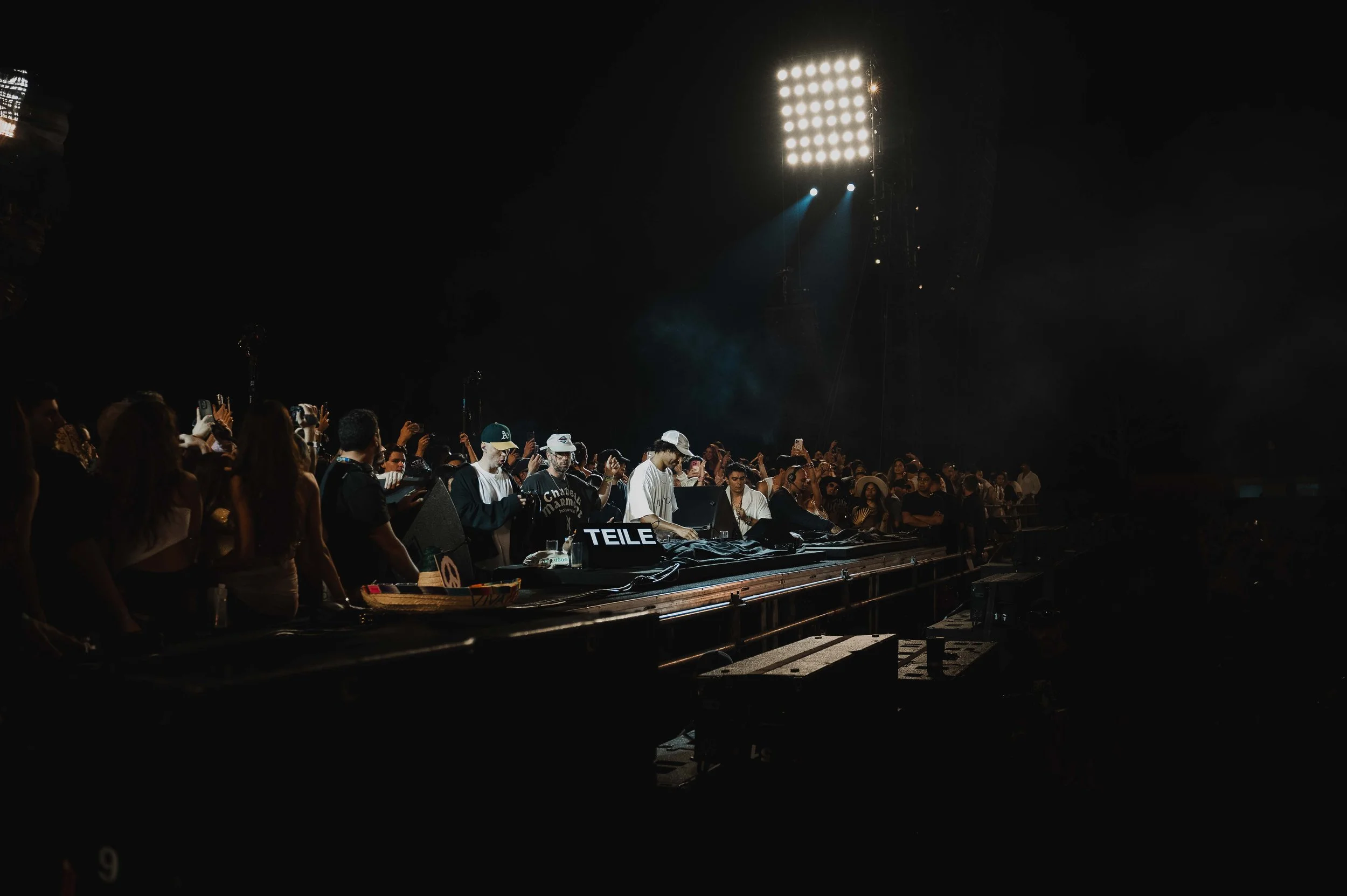 Crowd of people gathered at an outdoor night concert or music festival, with a DJ performing on stage and bright stage lights illuminating the scene.