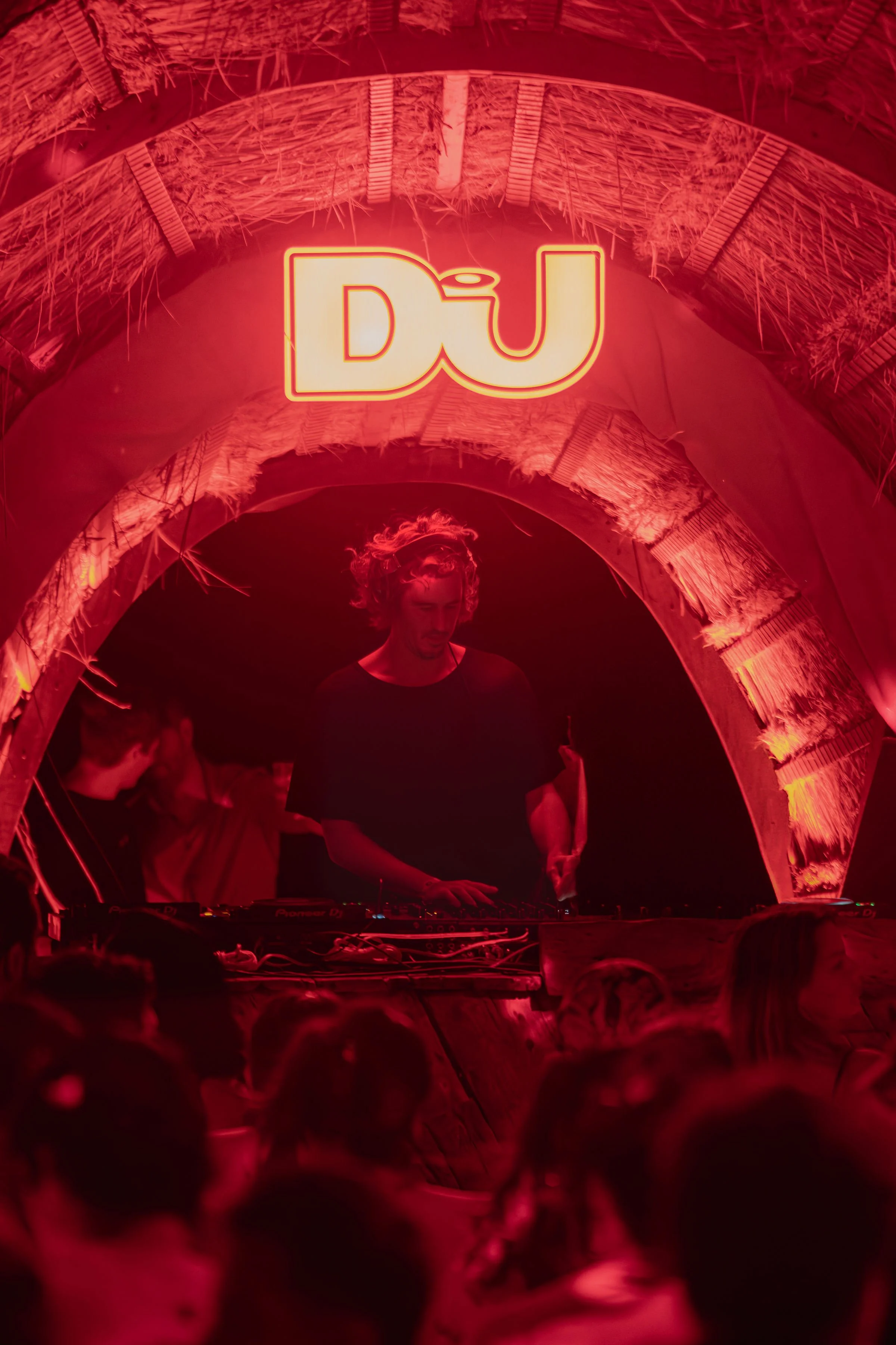 A DJ performing at a party under a red-lit, thatched arch with a neon sign displaying 'DØ' overhead, surrounded by a crowd of people.