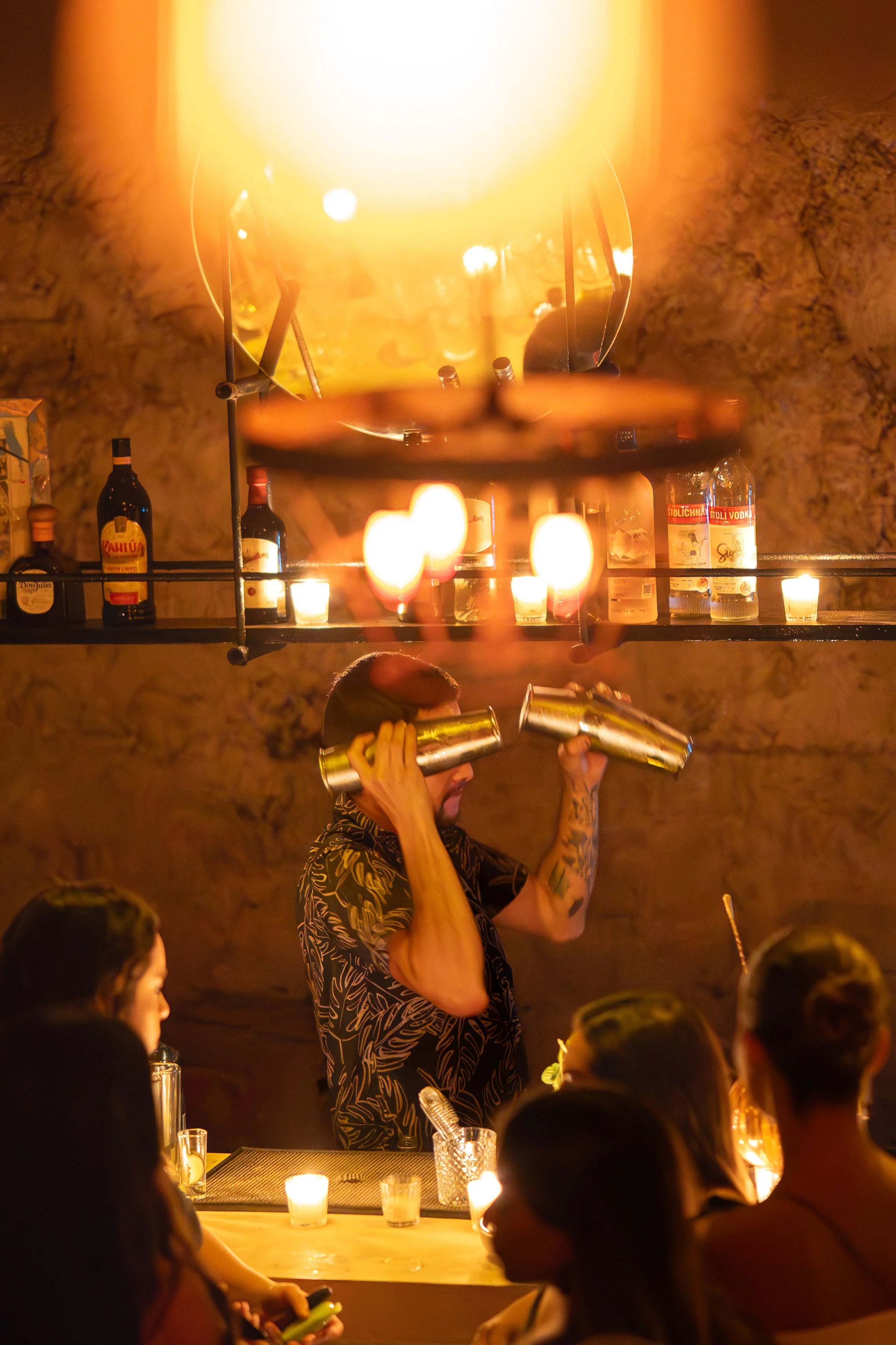 A bartender with tattoos and a tropical shirt, holding two cocktail shakers, stands behind a bar in a dimly lit setting, with a rocky wall, bottles, candles, and a large flame from a torch or flare above.