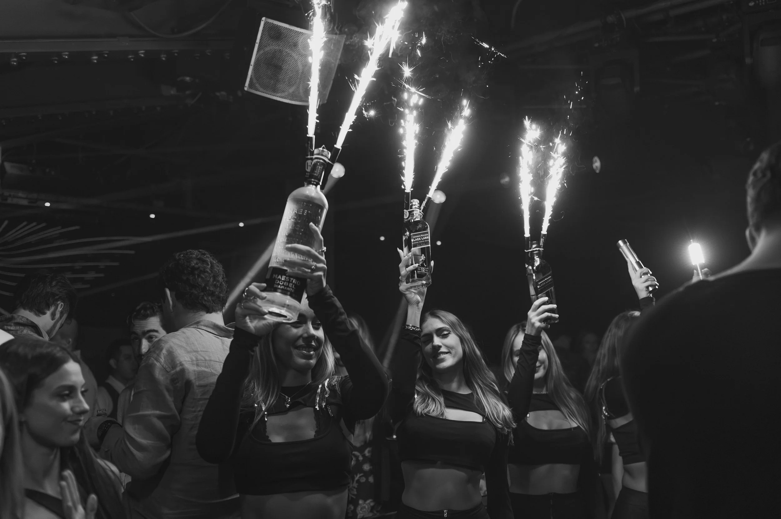 People celebrating at a party, holding bottles with sparklers attached, in a nightclub or party setting.