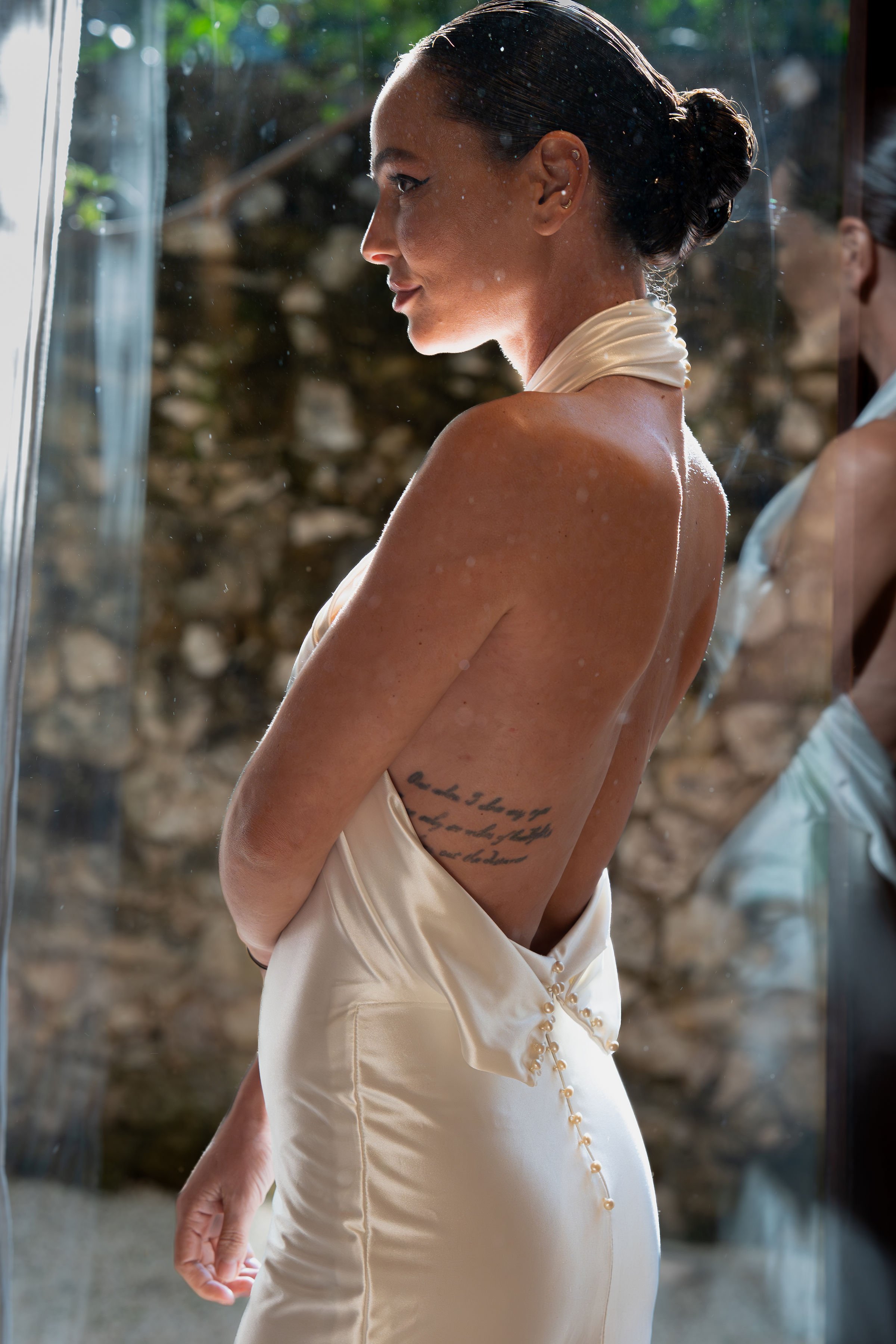 A woman with dark hair styled in an elegant bun is standing in front of a mirror, wearing a silky, off-shoulder dress with buttons on the back. She has a tattoo on her side visible through the open back of her dress. The background shows a stone wall