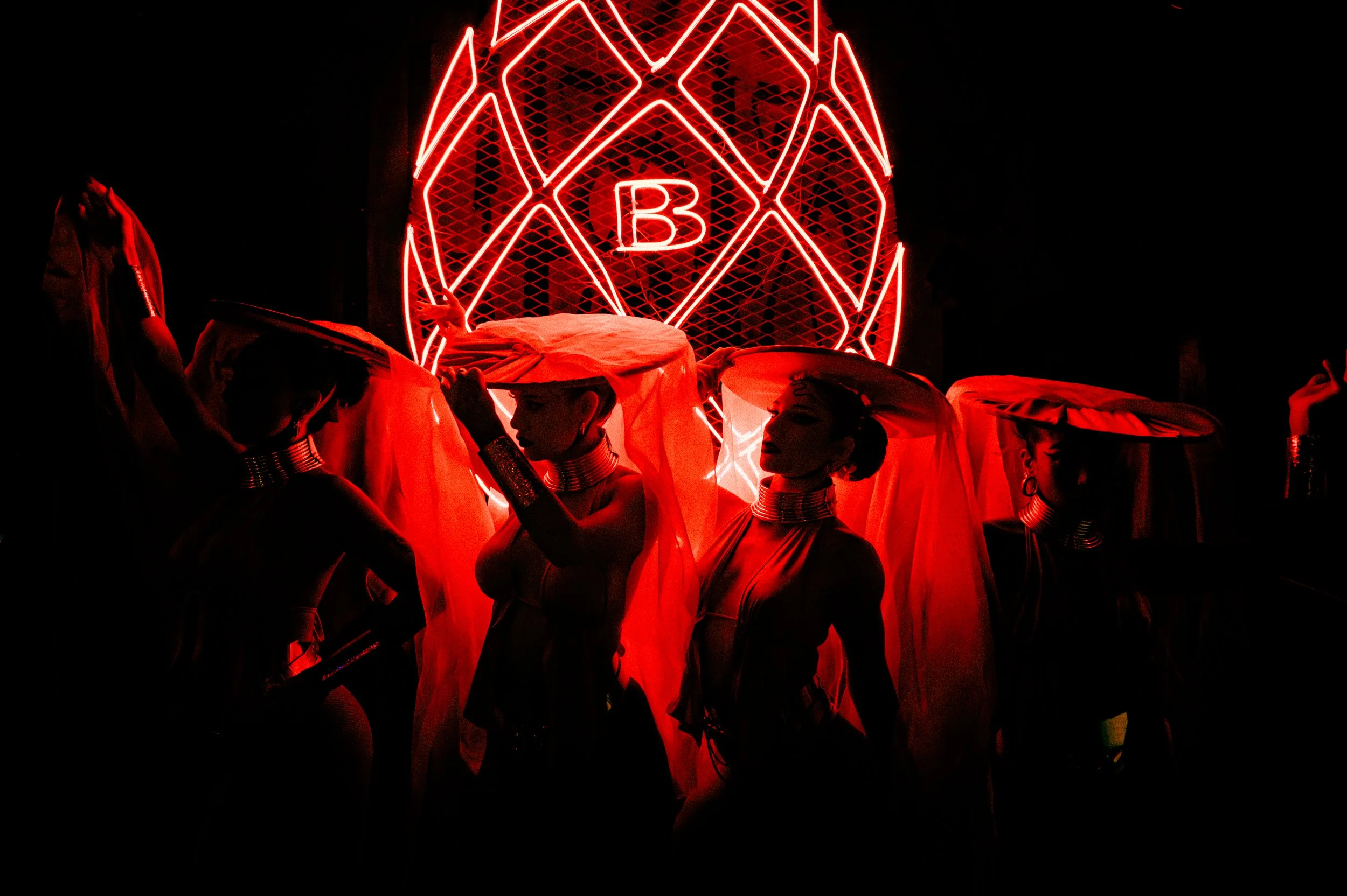 Silhouettes of women wearing traditional Egyptian-style costumes with headpieces and jewelry, standing in front of a red neon geometric design with the letter B at the center, in a dark setting.