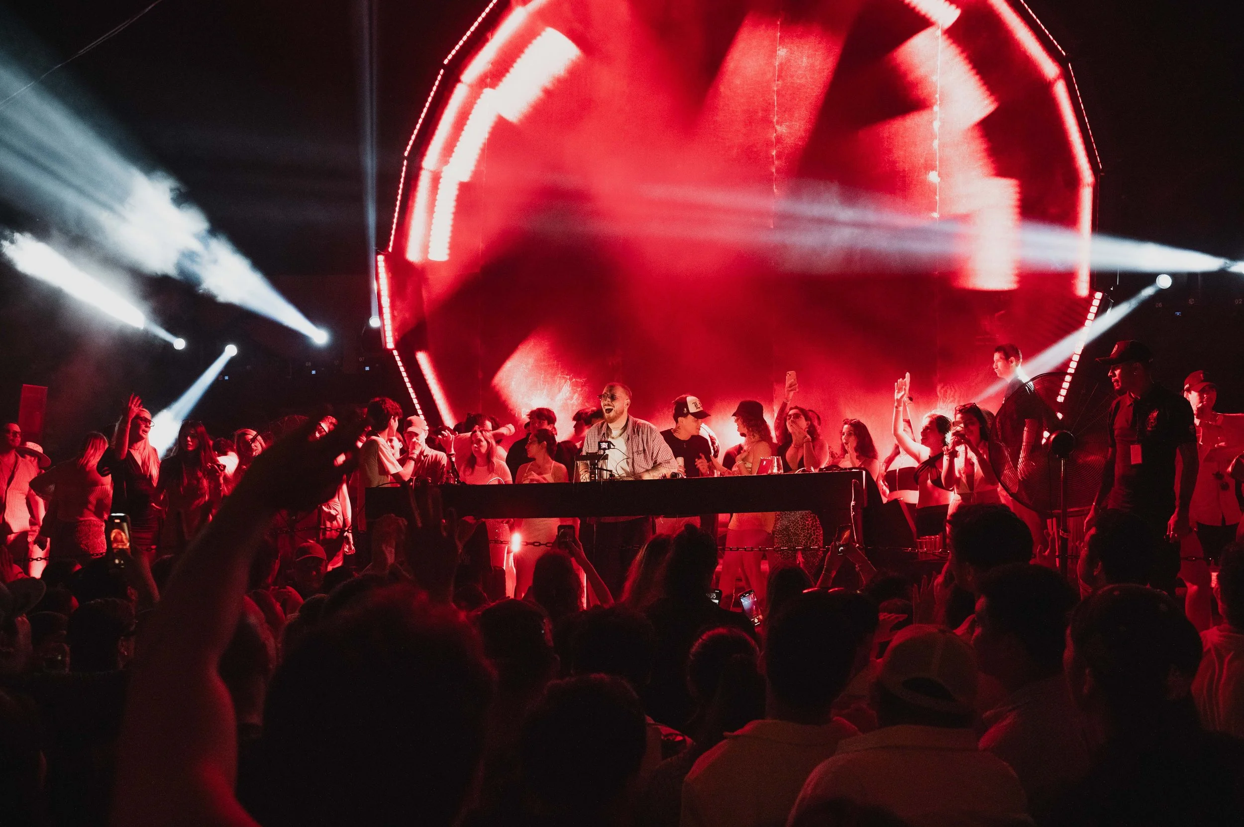 A DJ performs on stage at a concert with a large crowd, red lighting, and bright white spotlights in the background.