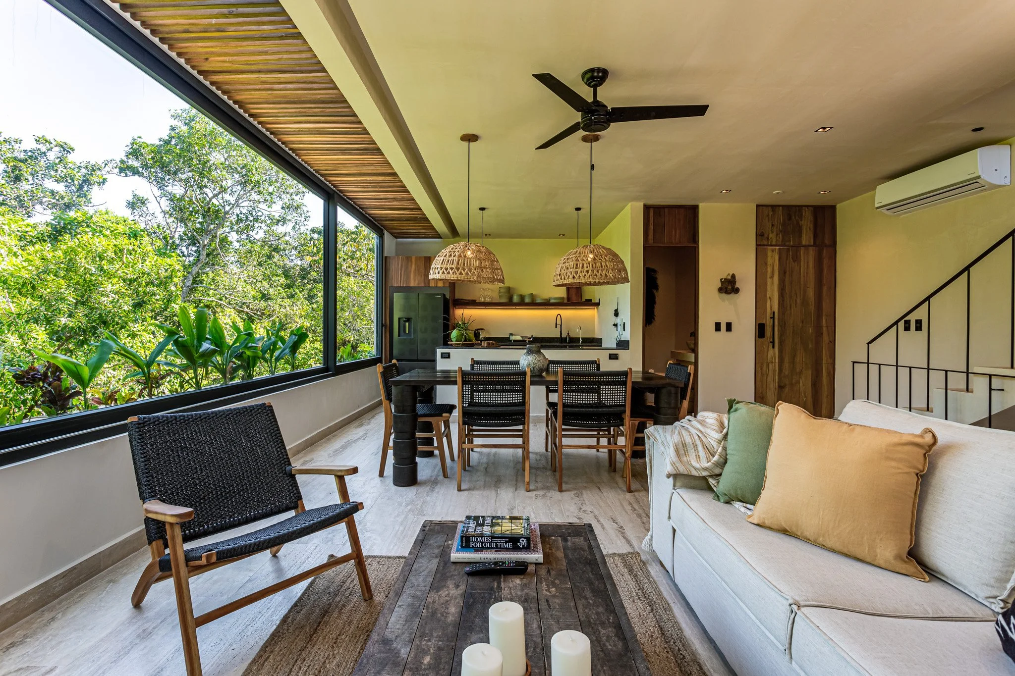 Living room with large window overlooking green trees, dining area with six chairs, standing lamp, white sofa with pastel pillows, wooden coffee table with candles and magazines, ceiling fan, air conditioner, staircase with black railing.