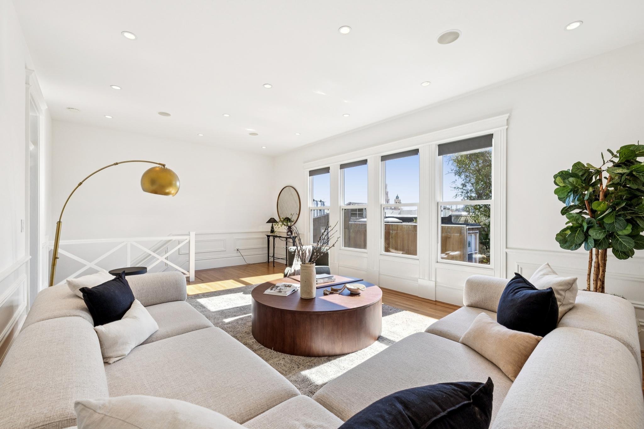 Bright living room with white walls and large windows, beige sectional sofa with black and white pillows, round wooden coffee table, decorative mirror, bookshelf, and large potted plant.
