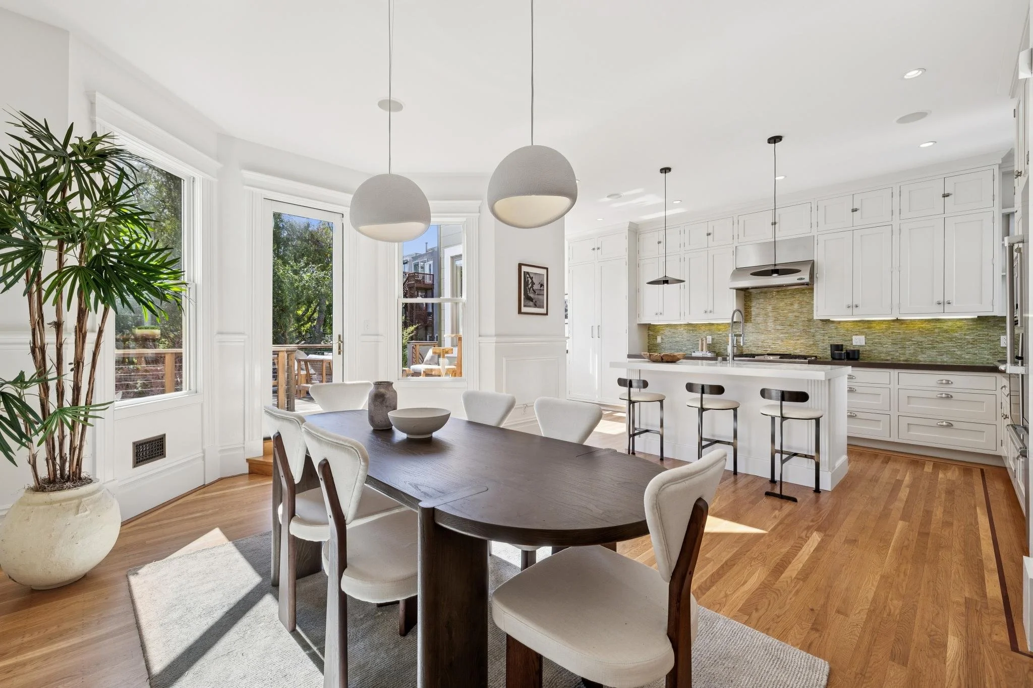 Bright kitchen and dining area with white cabinetry, a dark wood dining table, and white chairs, featuring large windows and pendant lighting.