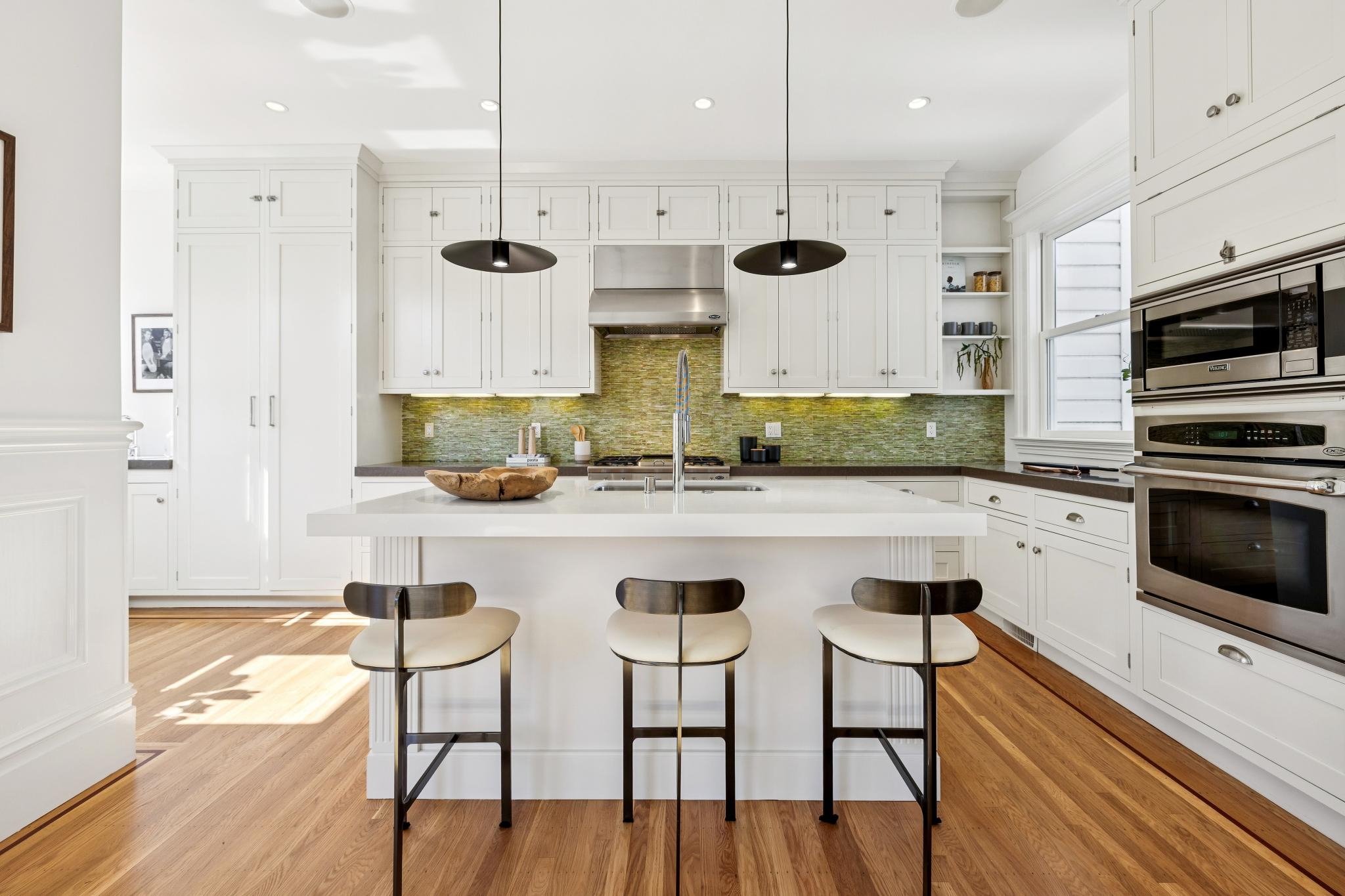 Modern kitchen with white cabinets, green backsplash, kitchen island, black bar stools, and stainless steel appliances.