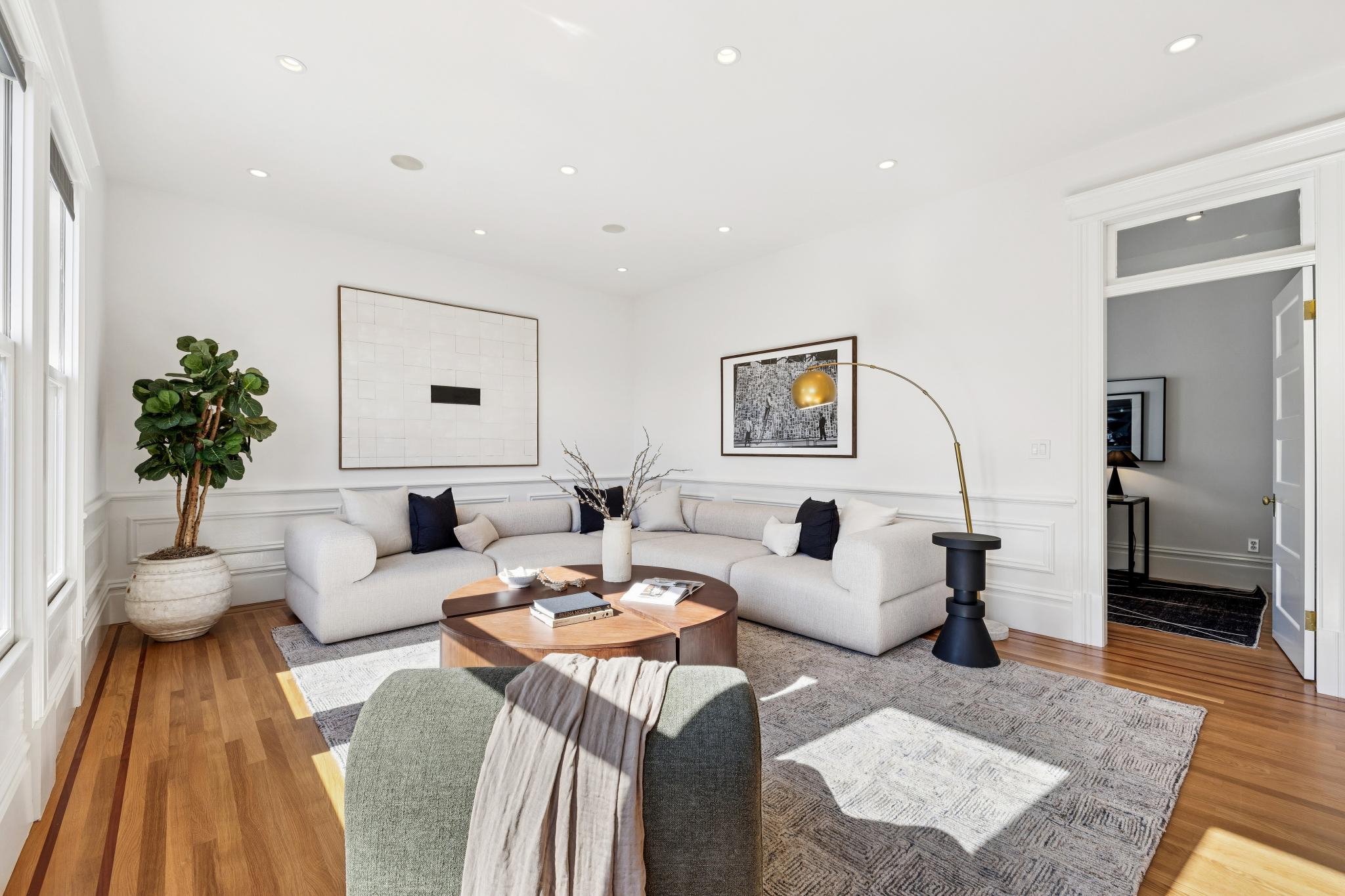 Living room with white walls, hardwood floors, a white curved sofa with black and white pillows, a round wooden coffee table, modern art on the walls, a large potted plant, a black side table, and a brass floor lamp.