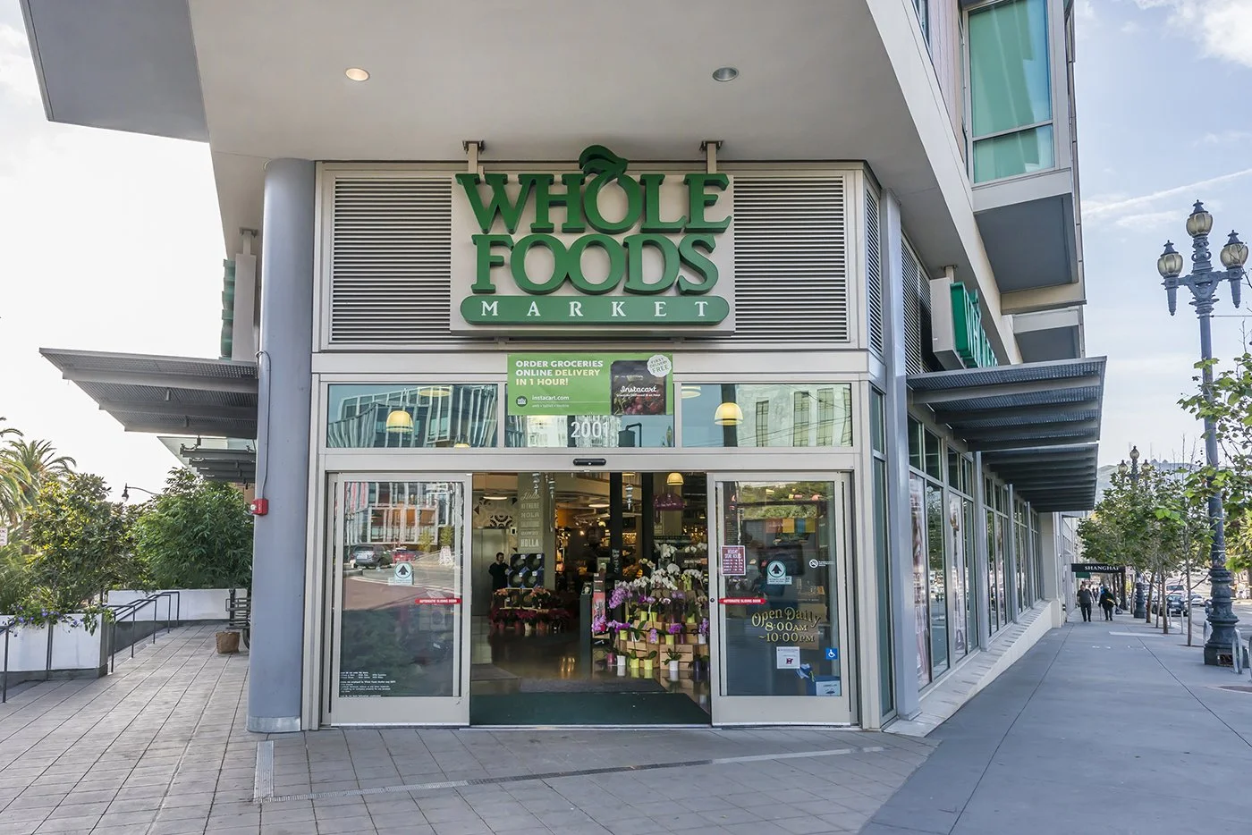 Exterior of Whole Foods Market store with glass doors, sign, and interior visible from outside. Sidewalk and street view.