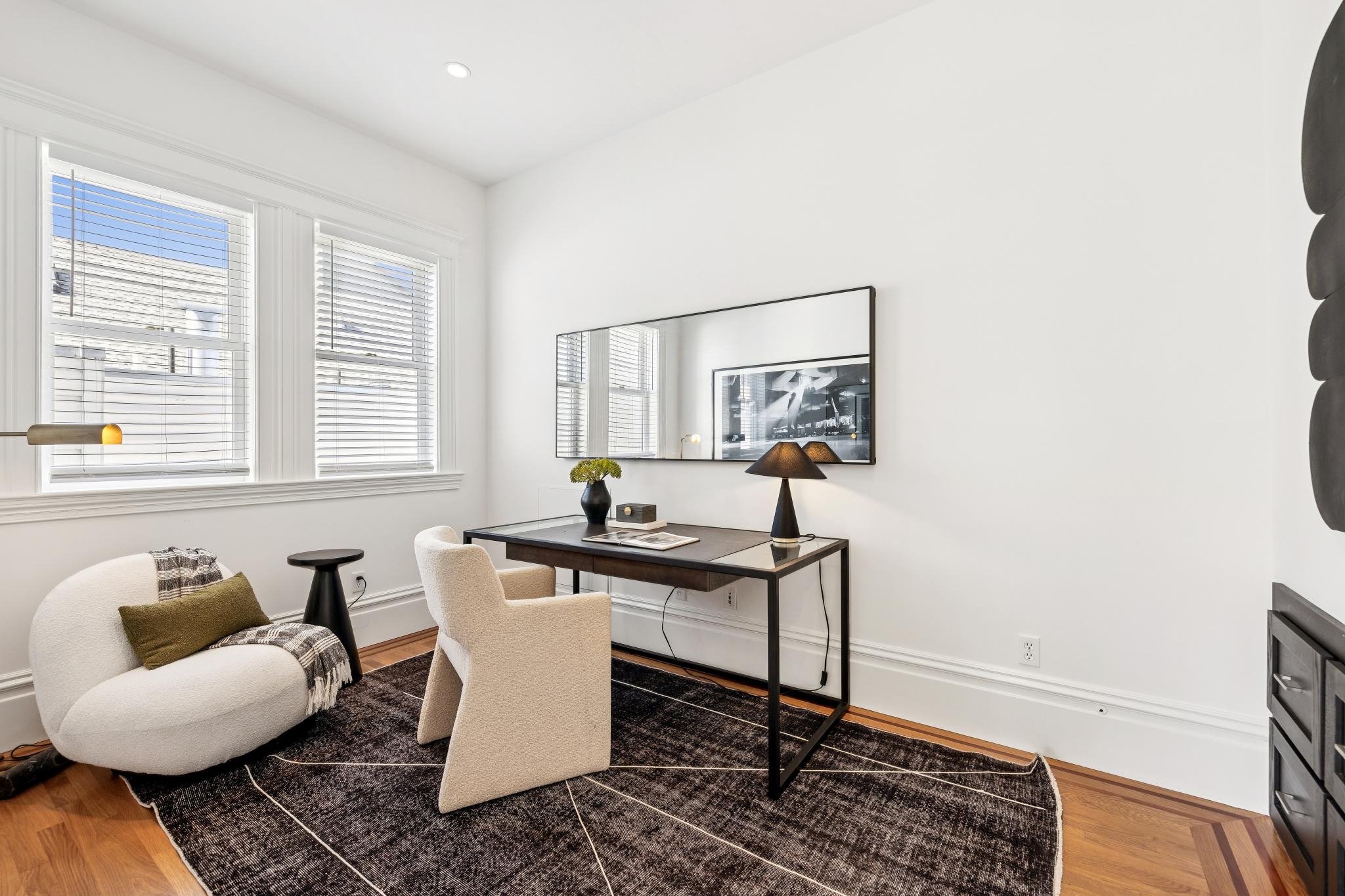 A modern, minimally decorated home office with three large windows, a black and wood desk, beige armchair, white oval chair with a brown pillow, a black lamp, and a large mirror on a white wall. There is a black and brown patterned rug on hardwood fl