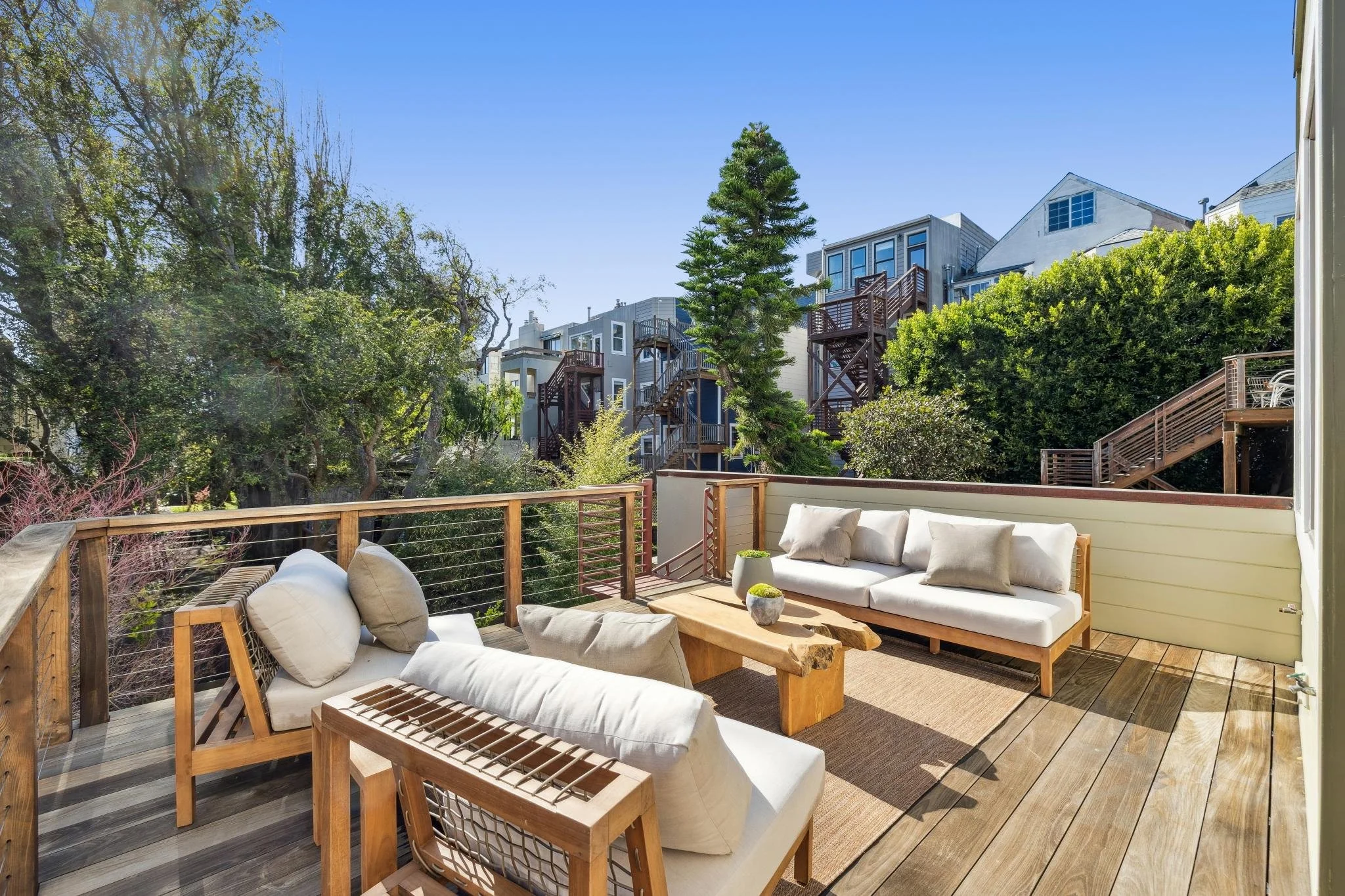 Outdoor patio with wooden deck, beige couches with cushions, a wooden coffee table, and potted plants, surrounded by lush green trees and residential buildings in the background.