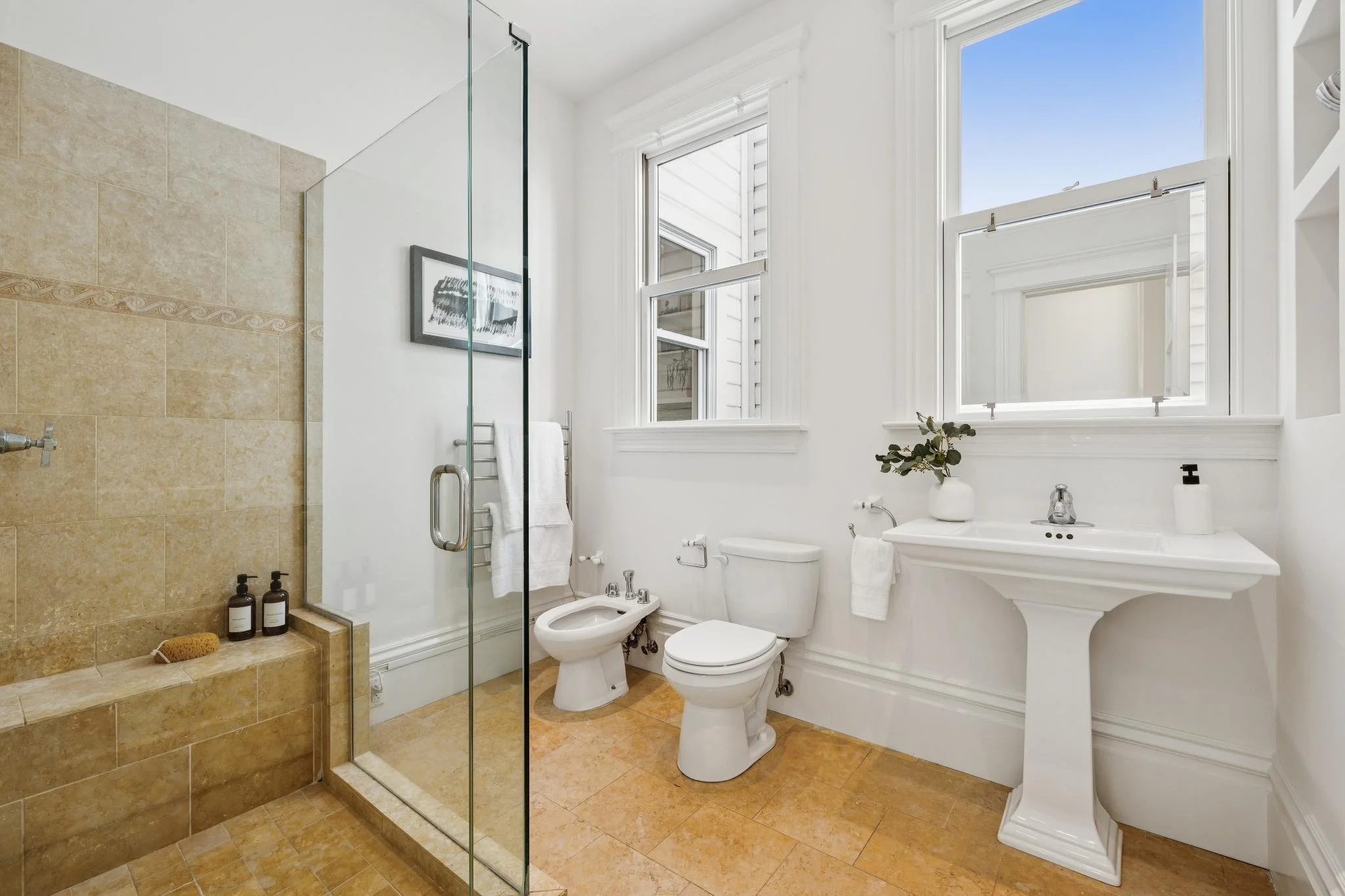 Bright bathroom with beige tile shower area, white toilet, bidet, and a pedestal sink. Large windows with white trim allow natural light; small potted plant and soap dispenser on sink, framed artwork on wall.