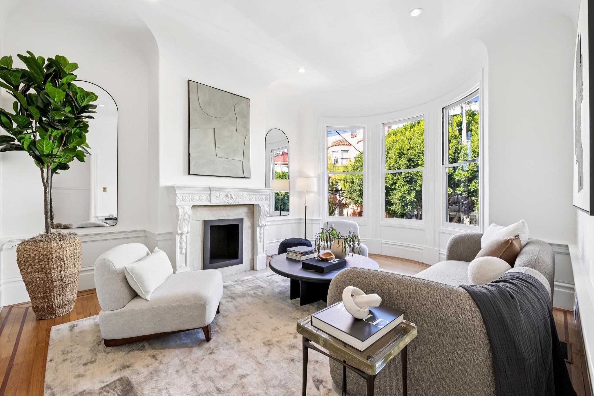 Bright living room with white walls and large bay windows, featuring a beige sofa with pillows, a white armchair, a round black coffee table with books and plants, a tall plant in a woven basket, a fireplace with a decorative mantel, and abstract art