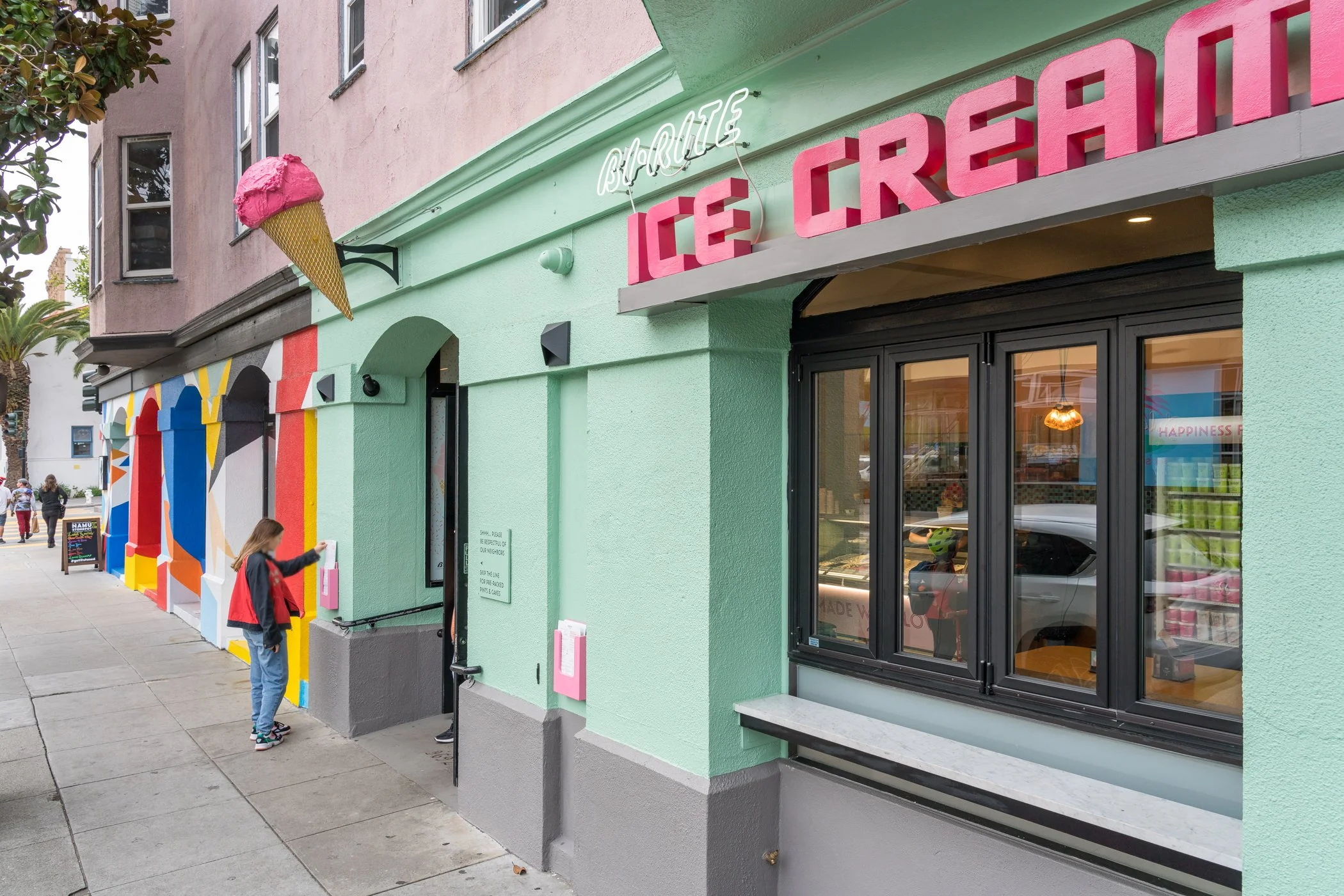 Colorful ice cream shop with pastel green exterior, large pink sign reading 'Ice Cream,' a 3D ice cream cone decoration, and a woman in red and black jacket ordering at the window. The building has a mural with abstract shapes along the sidewalk, and