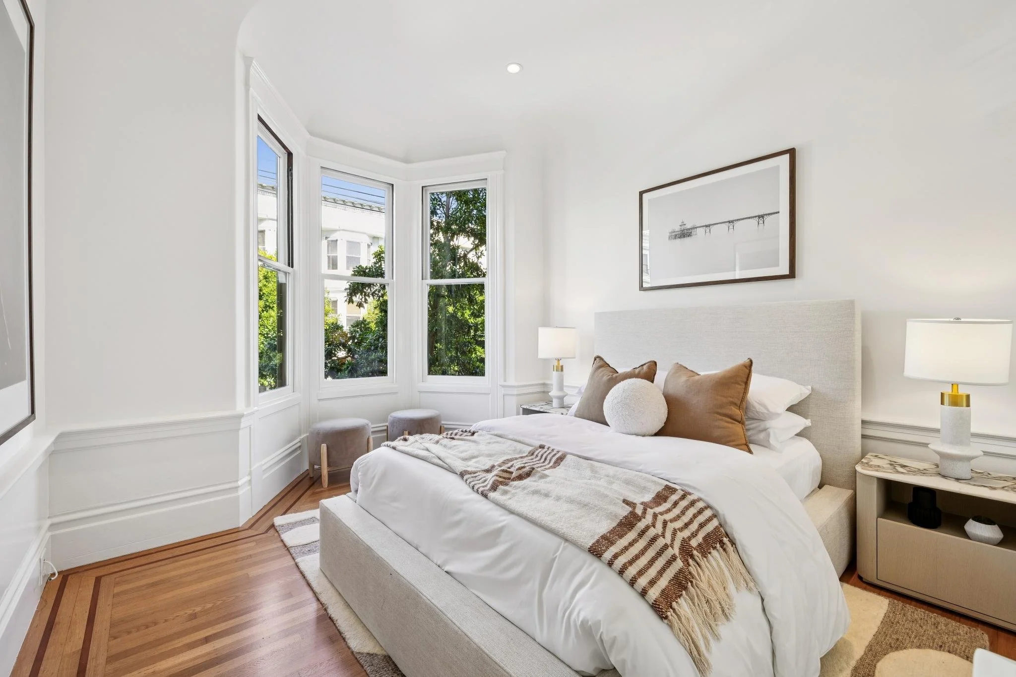Bright bedroom with white walls, large bay window, beige bed with white bedding, and decorative pillows. Nightstands with lamps on each side of the bed, wall art above headboard, and hardwood floors.