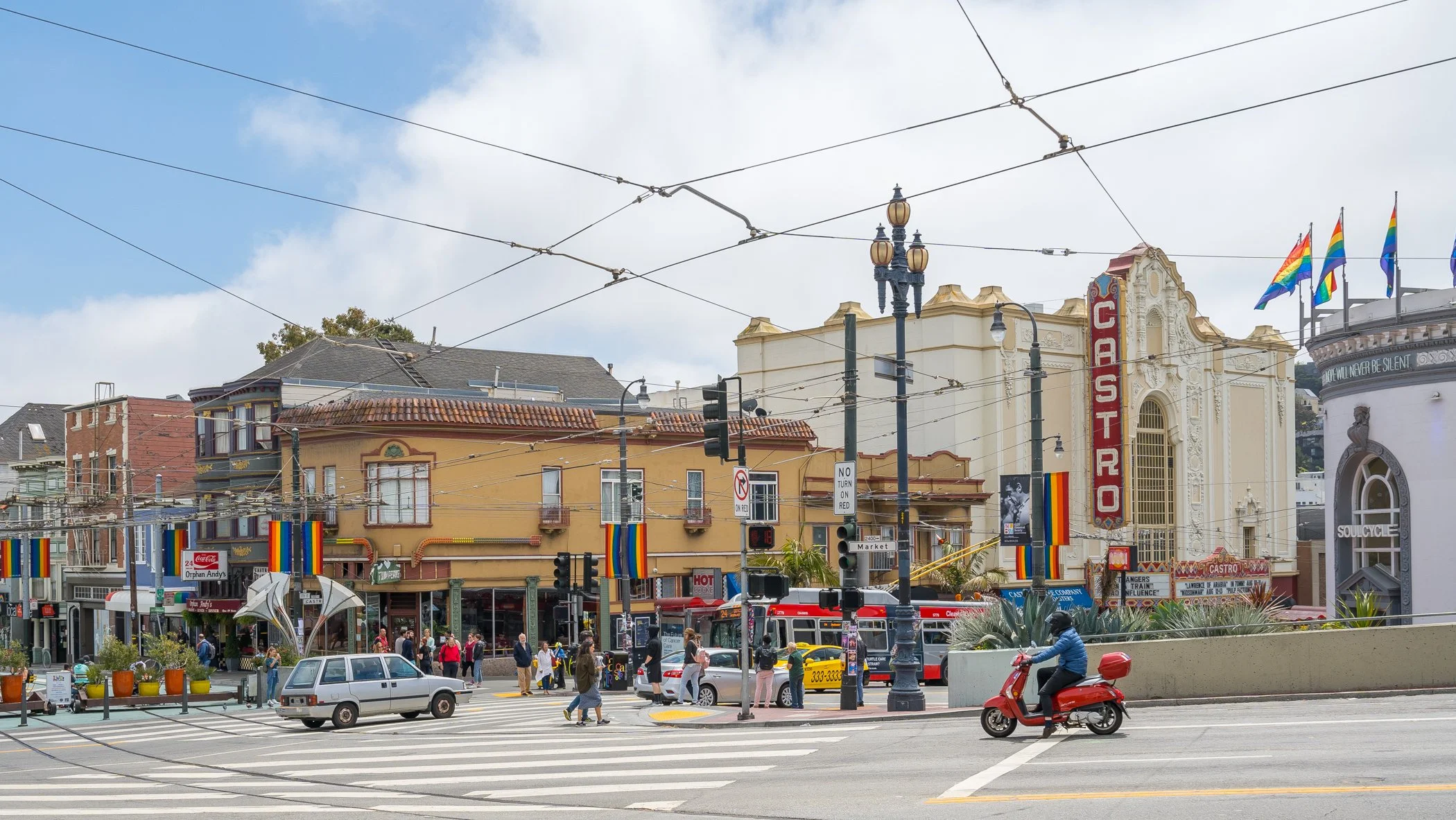 Street scene in San Francisco with pedestrians, cars, a red scooter, historic buildings, and rainbow flags. The Castro Theatre sign is visible.