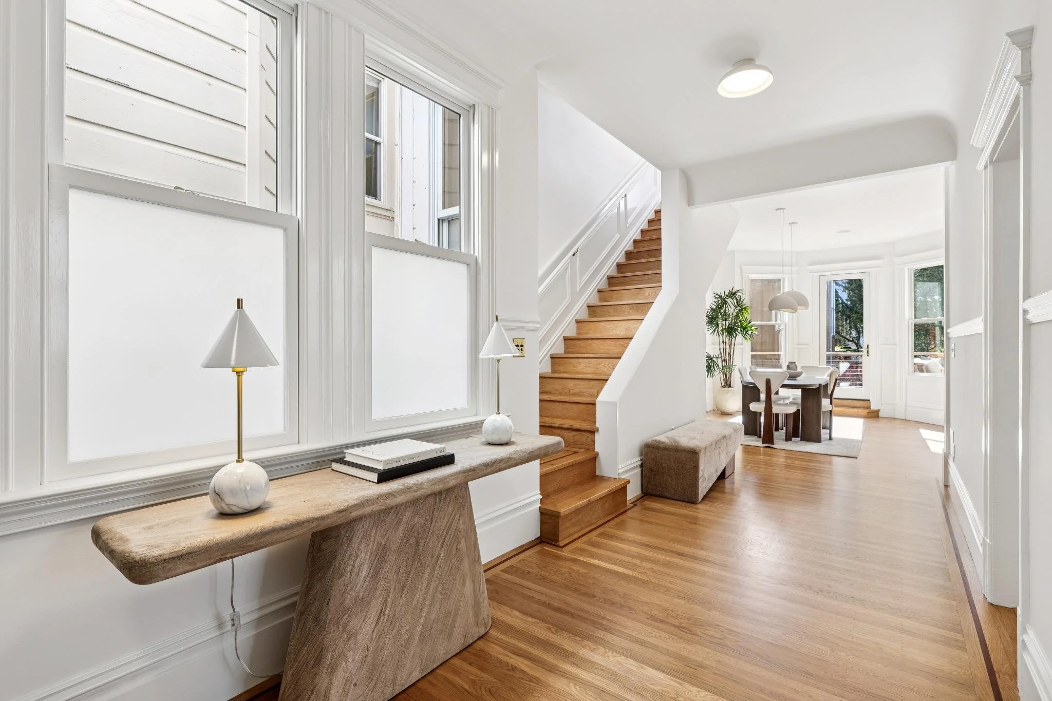 Bright and airy home's interior with wood flooring, white walls, large windows, a staircase, and a dining area with a round table and chairs.