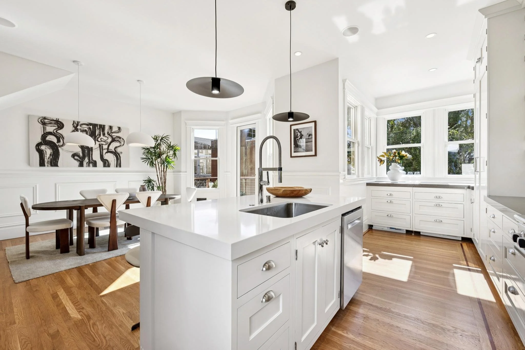 Bright white kitchen with large windows, white cabinets, and a central island with a sink, hardwood floor, dining area with table and chairs, and decorative artwork on the walls.