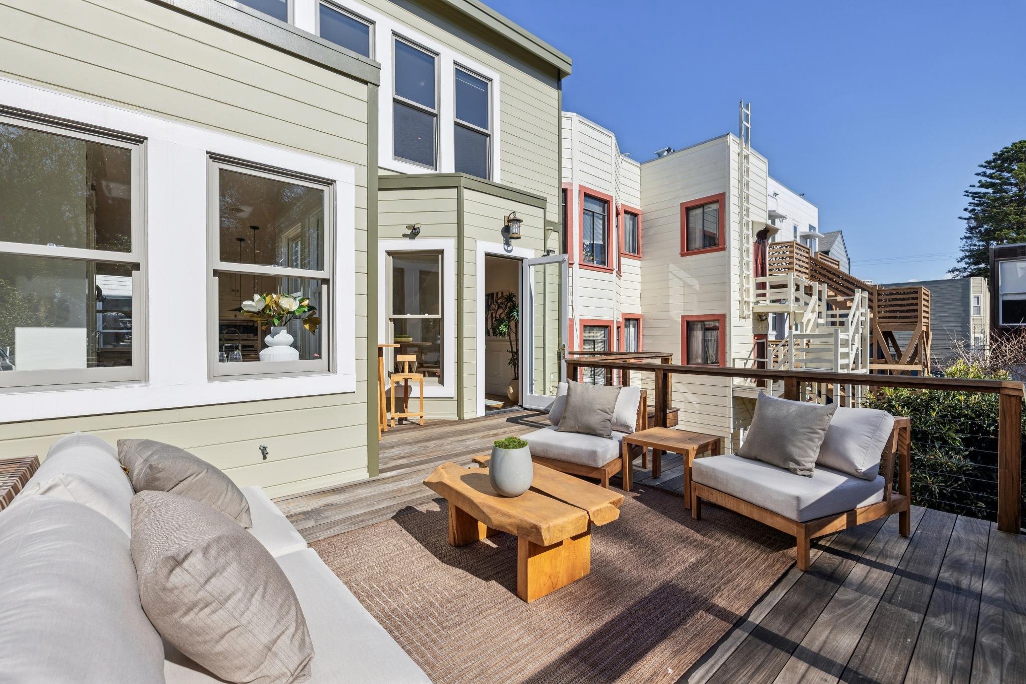 An outdoor balcony with wooden flooring, outdoor furniture including a white cushioned sofa and two armchairs, a small wooden table, and potted plants, with residential buildings in the background under a blue sky.