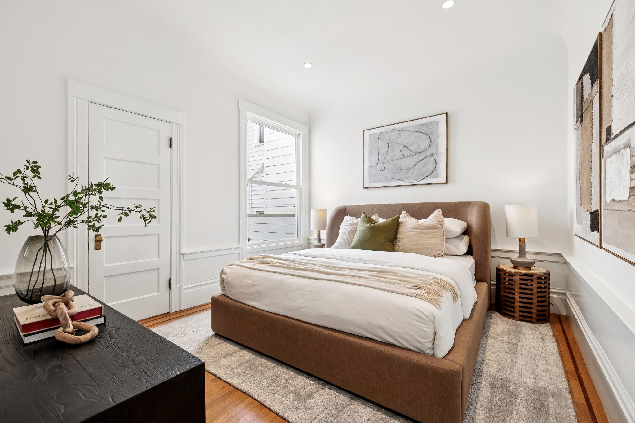A bedroom with a large bed with beige headboard, white bedding, multiple pillows, and a throw blanket. There is a window to the left of the bed, a dark wooden nightstand with a lamp on each side, abstract artwork above the bed, a plant on a black dre