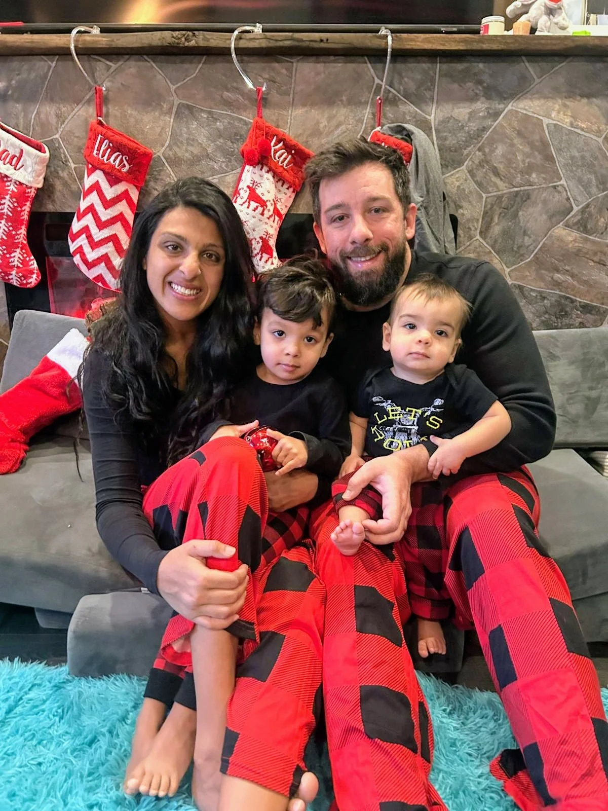 A family of four sitting on a couch in front of a fireplace decorated with Christmas stockings. The family members are smiling and wearing matching red plaid pajamas. The mother has long dark hair, and the father has a beard. The two children, a boy and a younger toddler, are sitting between their parents. There is a Christmas stocking with the name "Lilas" and others with names embroidered on them hanging above the fireplace.