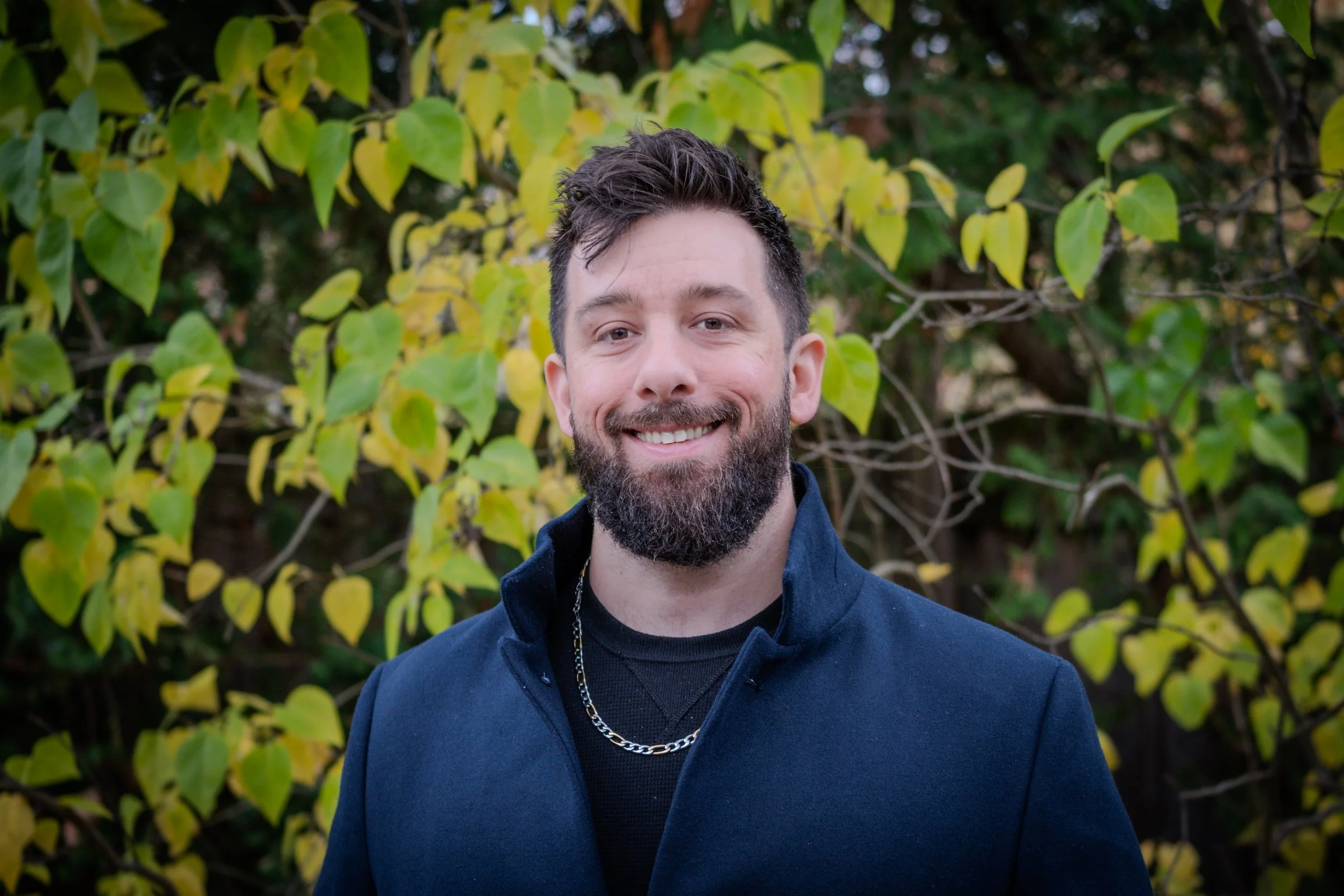 A smiling man with dark hair and a beard, wearing a navy blue jacket and a silver chain, standing outdoors in front of green and yellow leafy bushes.