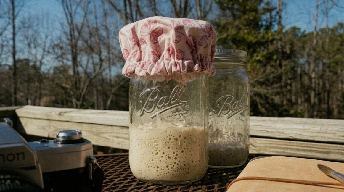 Two mason jars, one covered with a cloth, sitting on a table outdoors. One jar contains a foamy beverage, and the other appears to contain a mixture or powder. A camera and a closed notebook are also on the table with a wooded background.