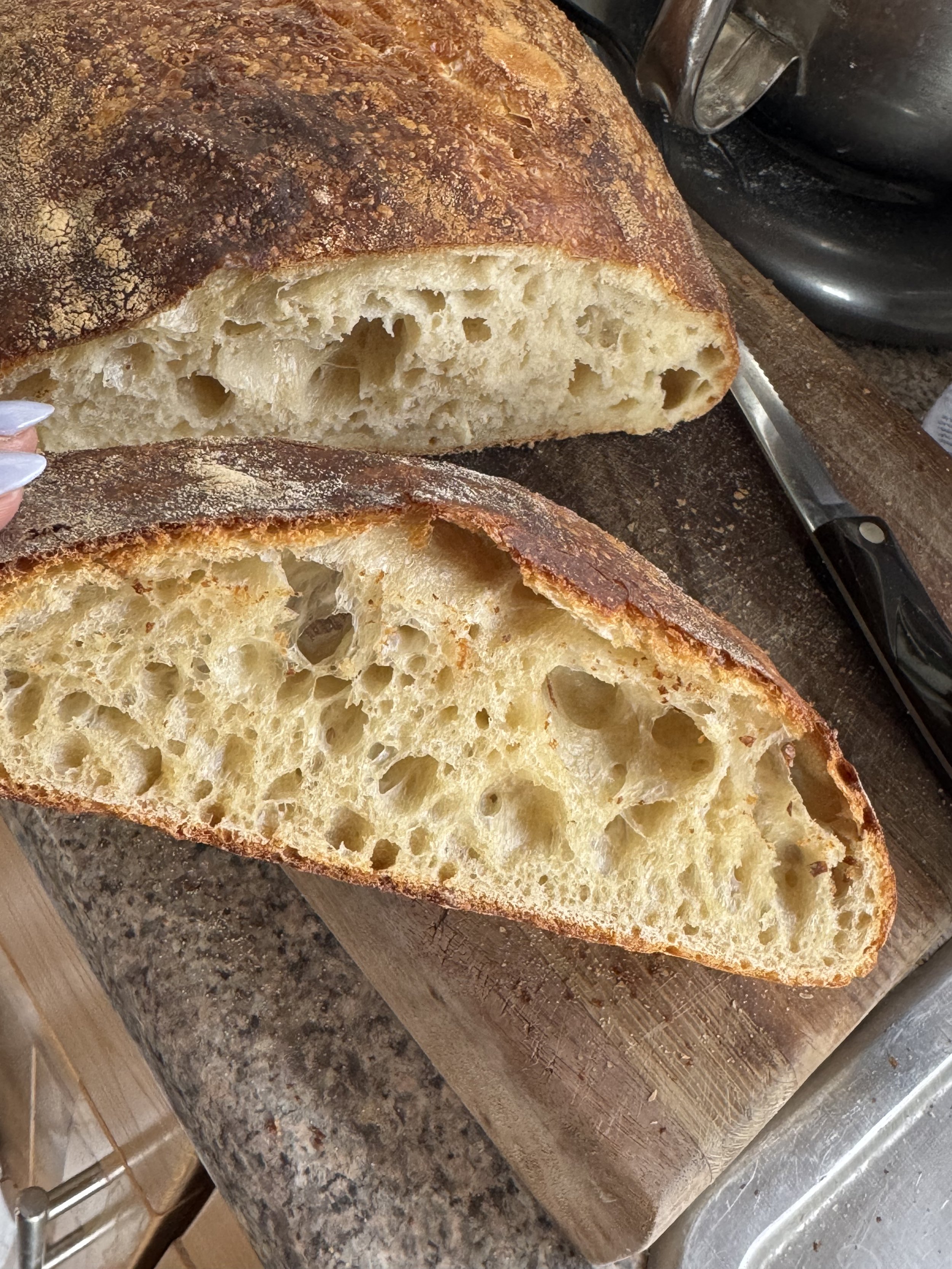 Close-up of a round loaf of bread with a crusty, browned exterior, sliced open to reveal a soft, airy, and bubbly interior on a wooden cutting board with a bread knife nearby.