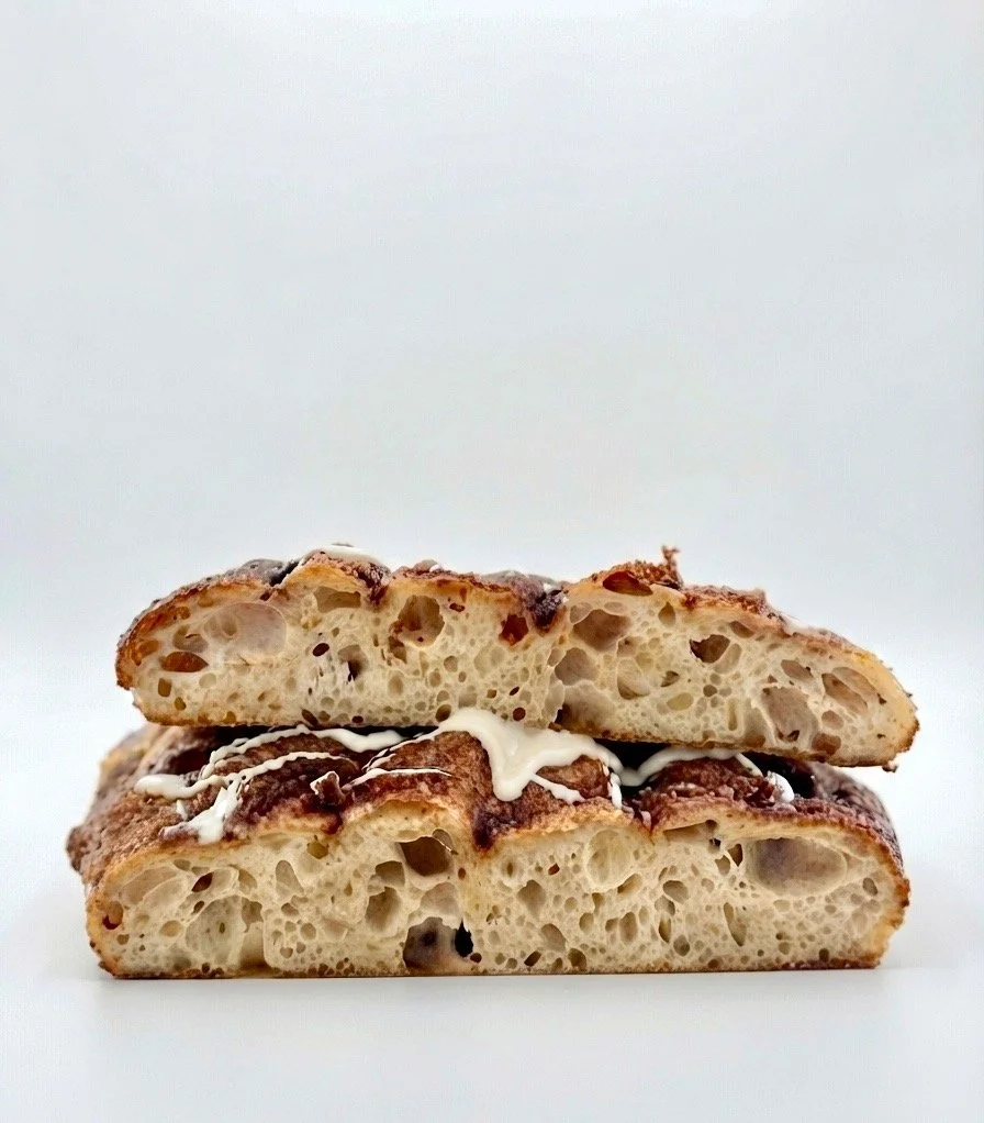 Close-up of two slices of focaccia bread with golden crust, airy interior, and visible olives, stacked on a white surface.